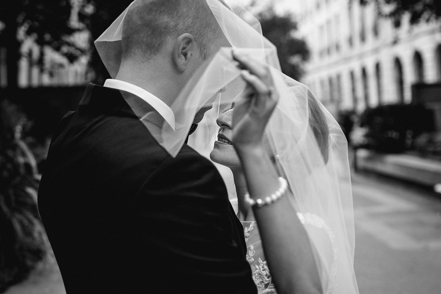 Black and white photo of newlyweds sharing an intimate moment on urban sidewalk with veil and formal attire.