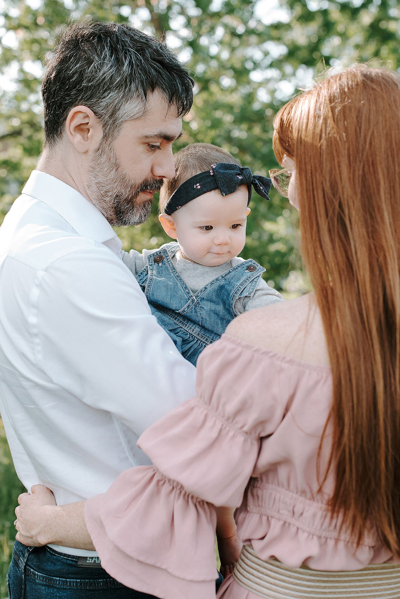 Family sharing tender moment outdoors with baby wearing denim overalls and black headband in natural setting.
