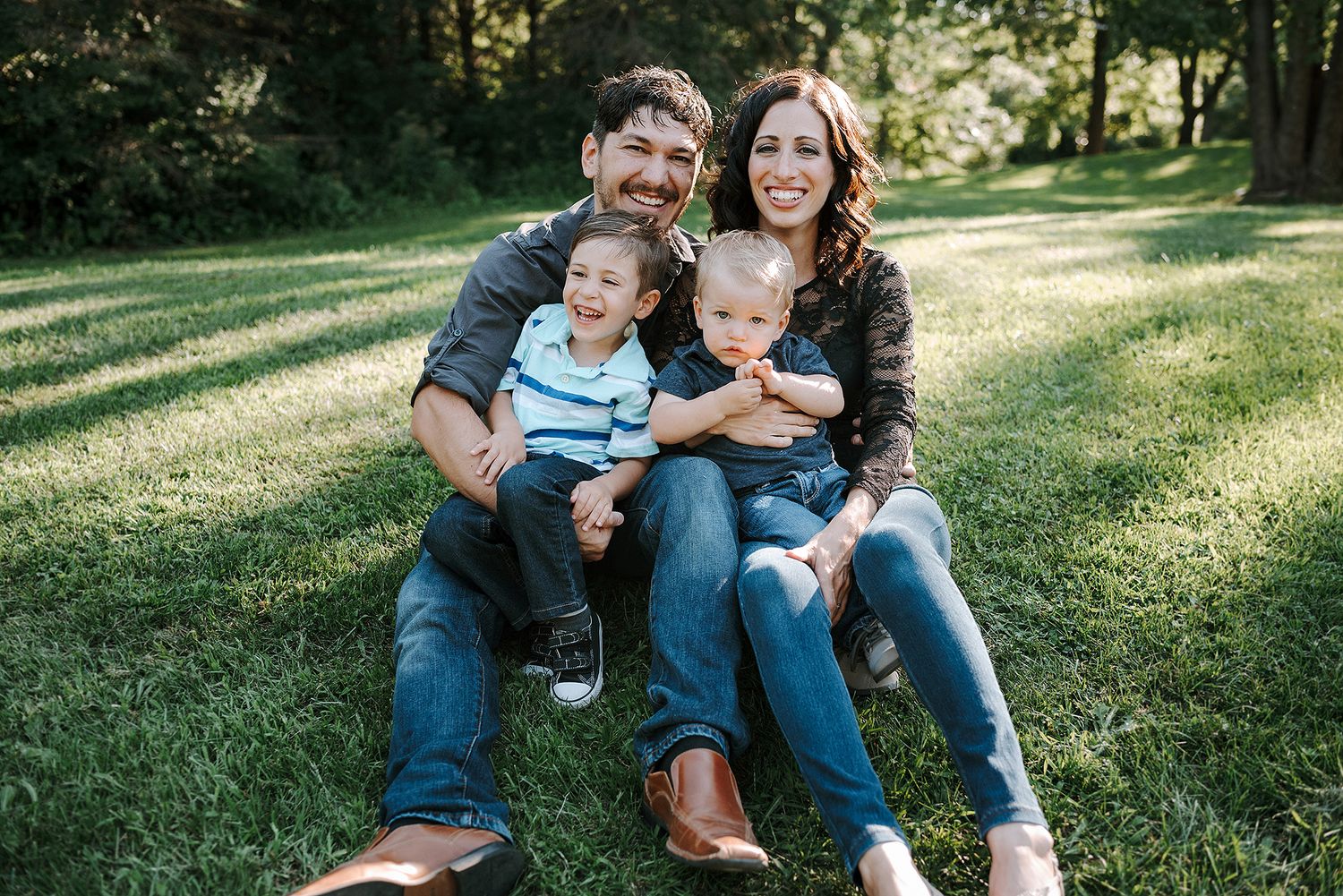 A family sits together on grass in a sunlit park during a casual outdoor photo session.