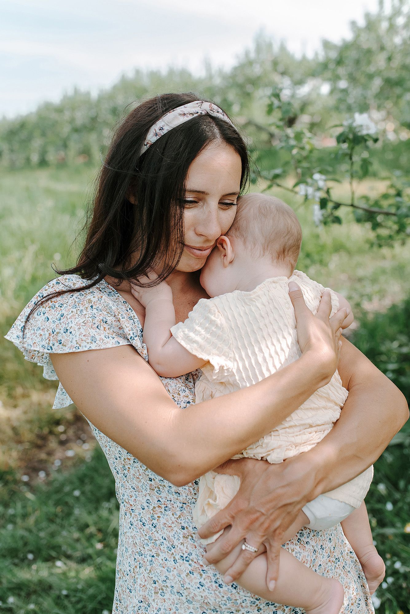 A loving parent in a floral dress holding and kissing a baby outside in a garden setting during summer.