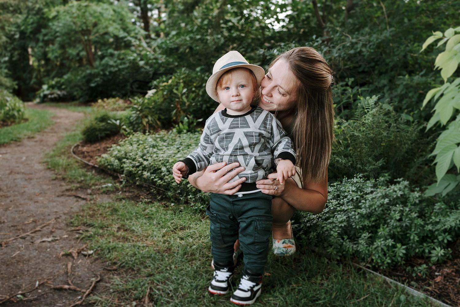 A parent and child share a tender moment while walking along a wooded garden path surrounded by lush greenery.