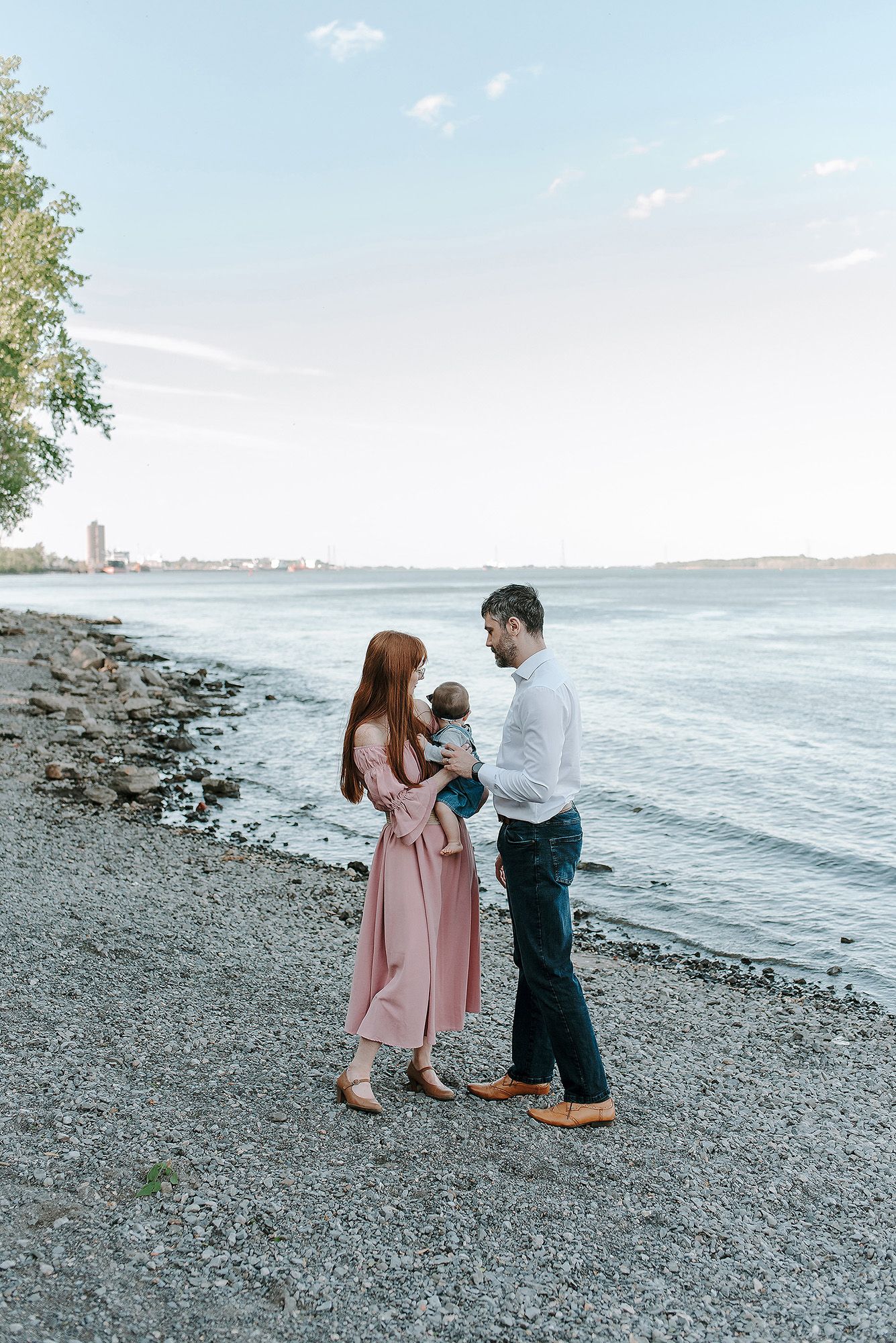 A couple stands together on a rocky beach shoreline at sunset with city buildings visible in the distance.