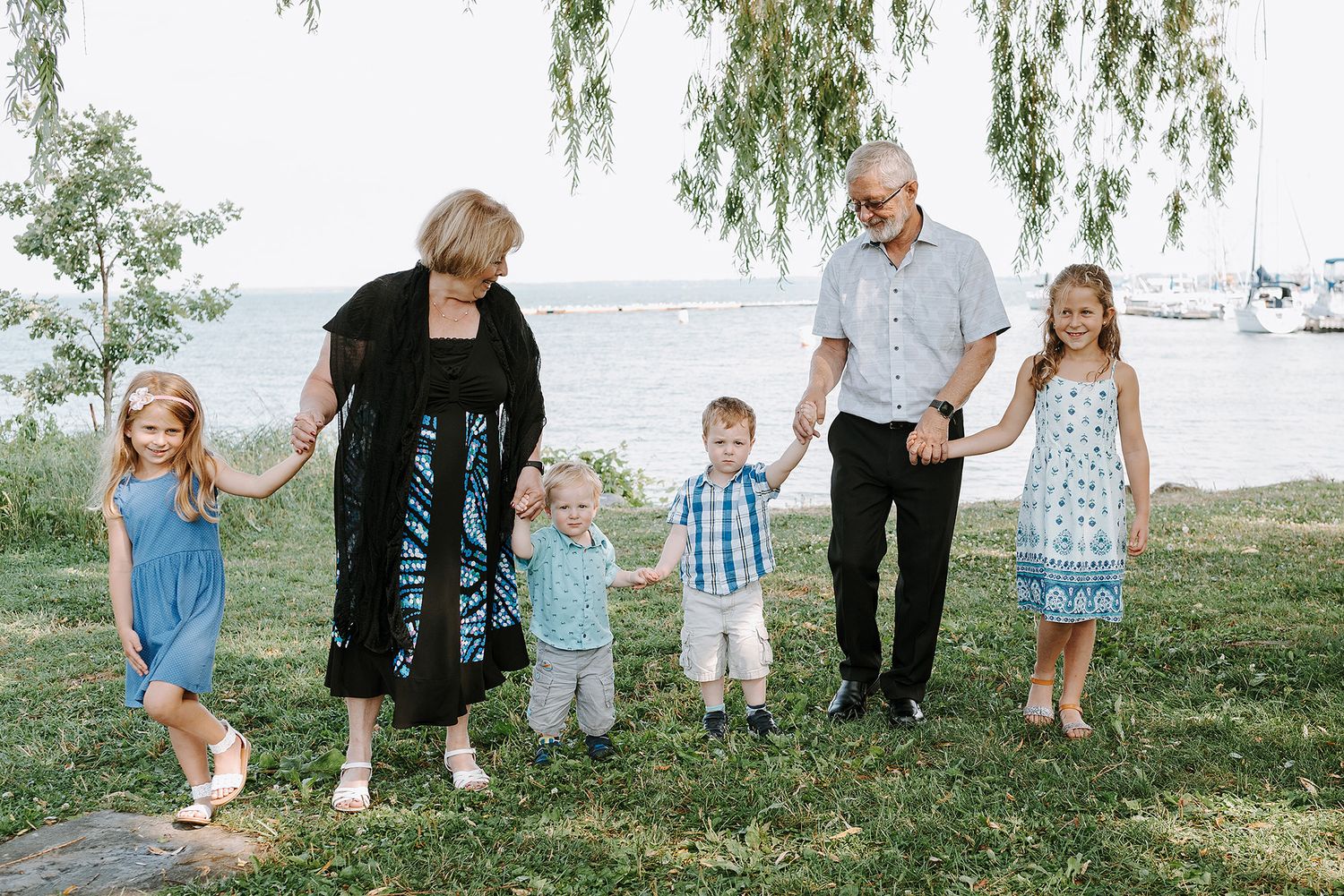 A family holds hands while standing together on grassy waterfront grounds under a weeping willow tree.