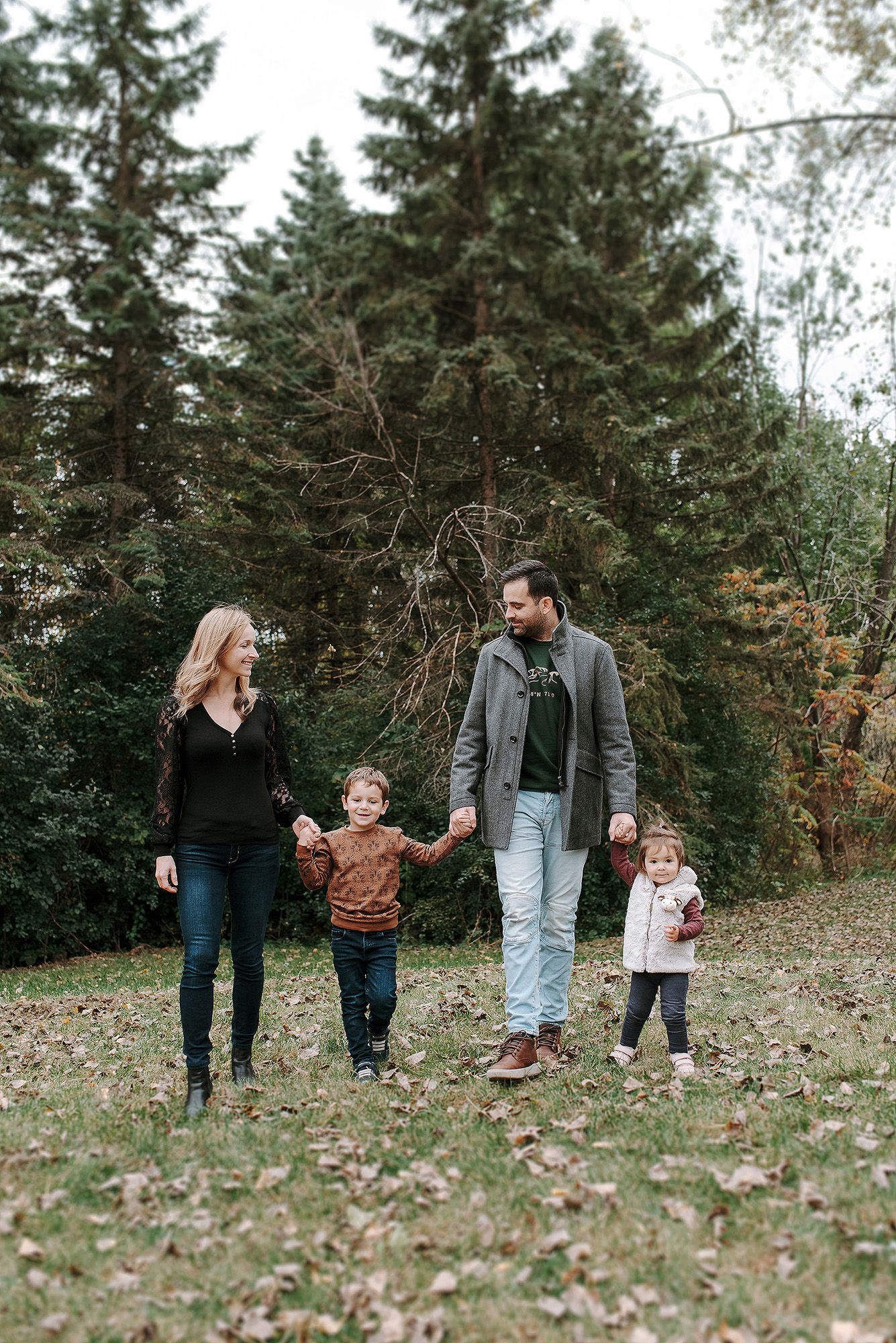 A family walks together holding hands along a wooded path surrounded by evergreen trees on an autumn day.