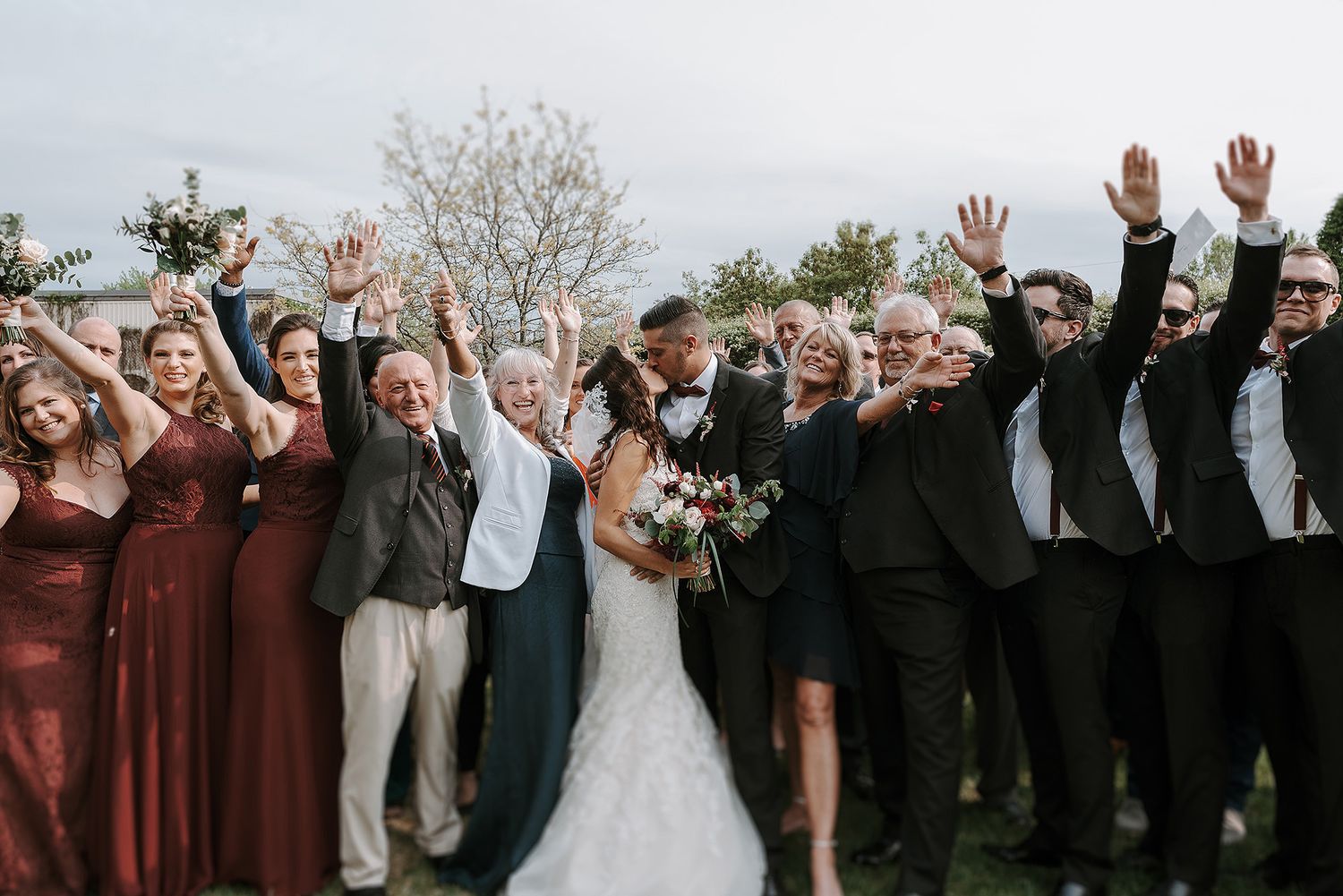 A wedding party raises their hands joyfully while posing together outdoors with bridesmaids in burgundy dresses. Espace Rive-Gauche.