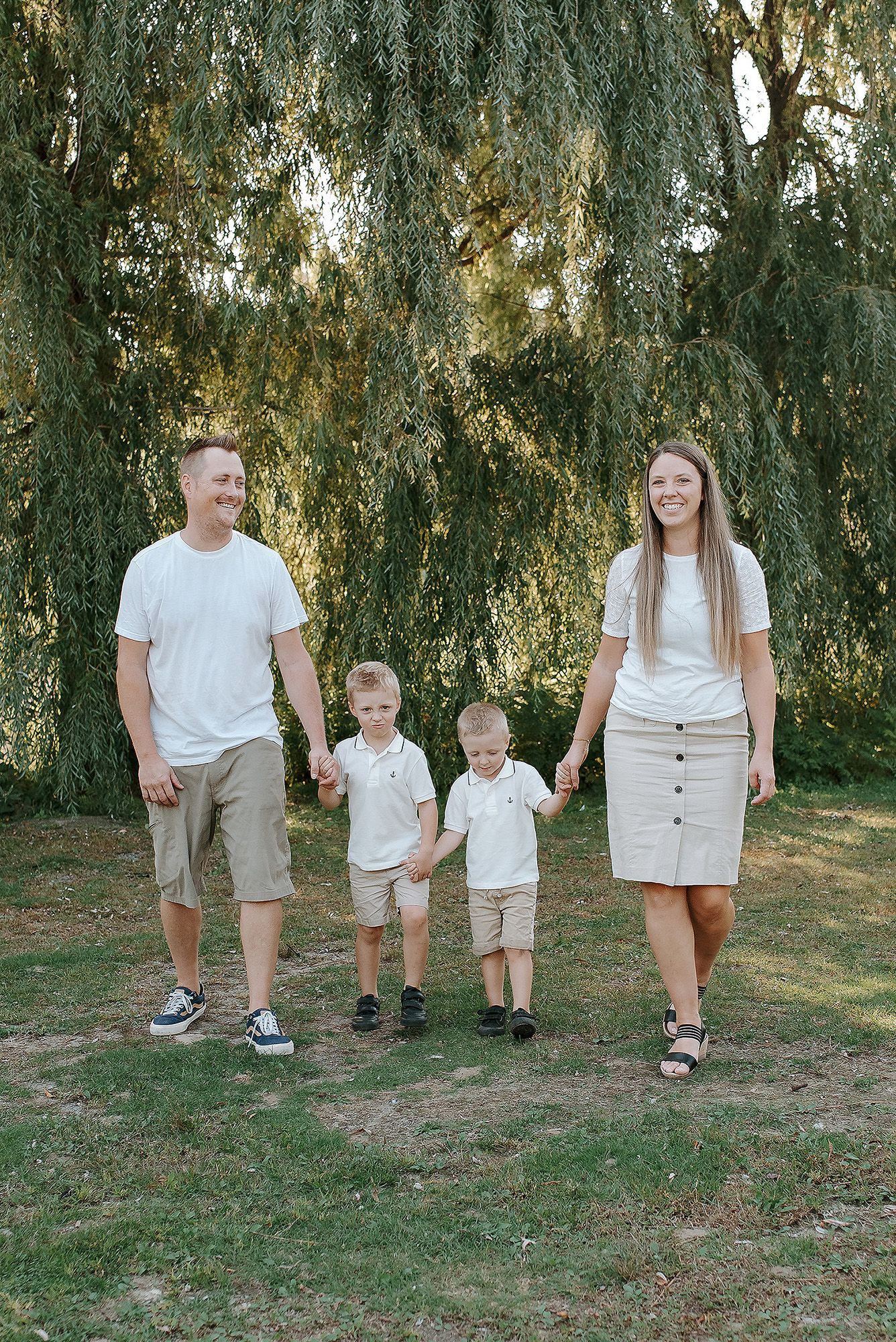 A family of four stands together in coordinated light-colored outfits beneath a weeping willow tree on a sunny day.