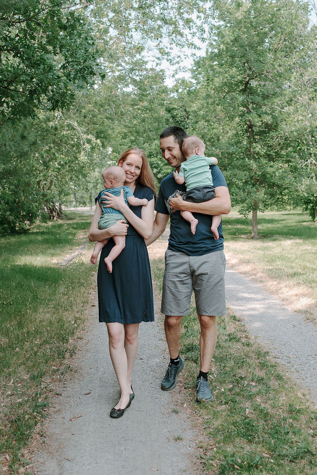 A couple walking on a path in a park holding children in matching teal outfits on a sunny summer day.