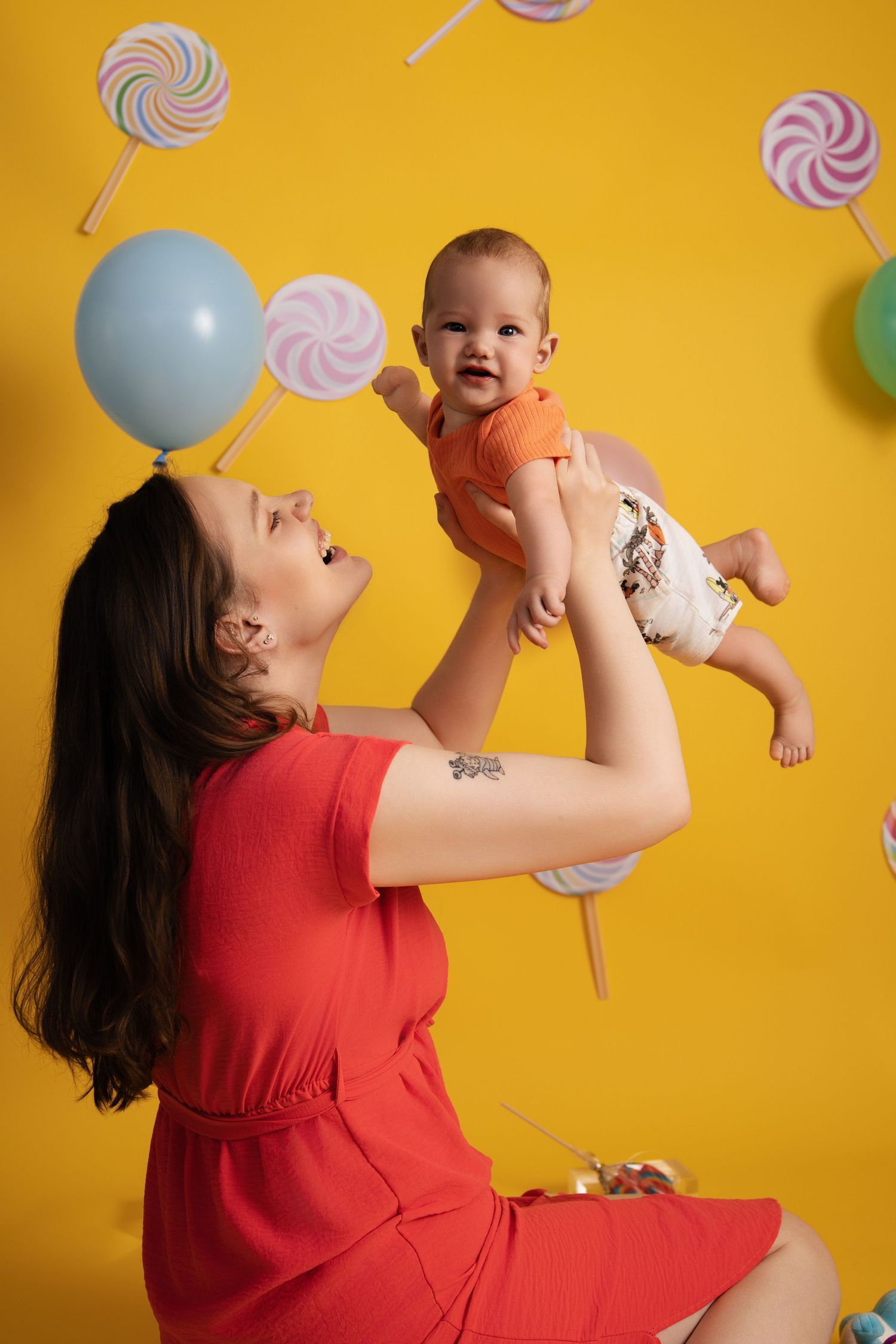 Woman in red dress playing with joyful baby against yellow wall with lollipop decorations in fun lifestyle photo series.