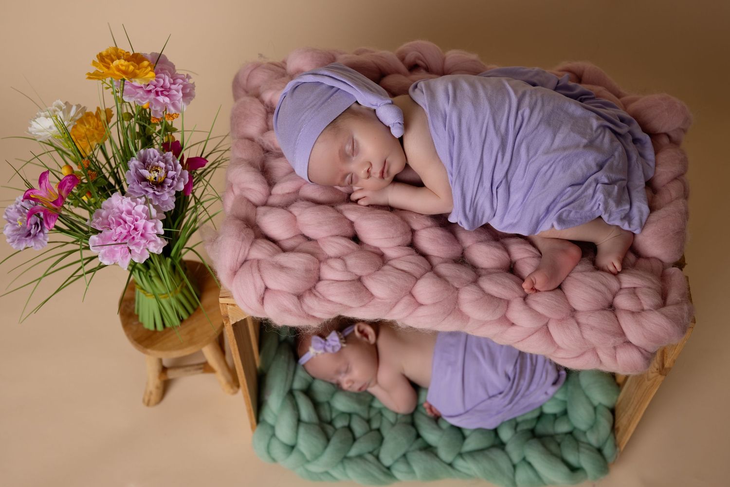 A sleeping baby lies on chunky knit blankets in pink and mint green, with a floral arrangement on a wooden stool nearby.