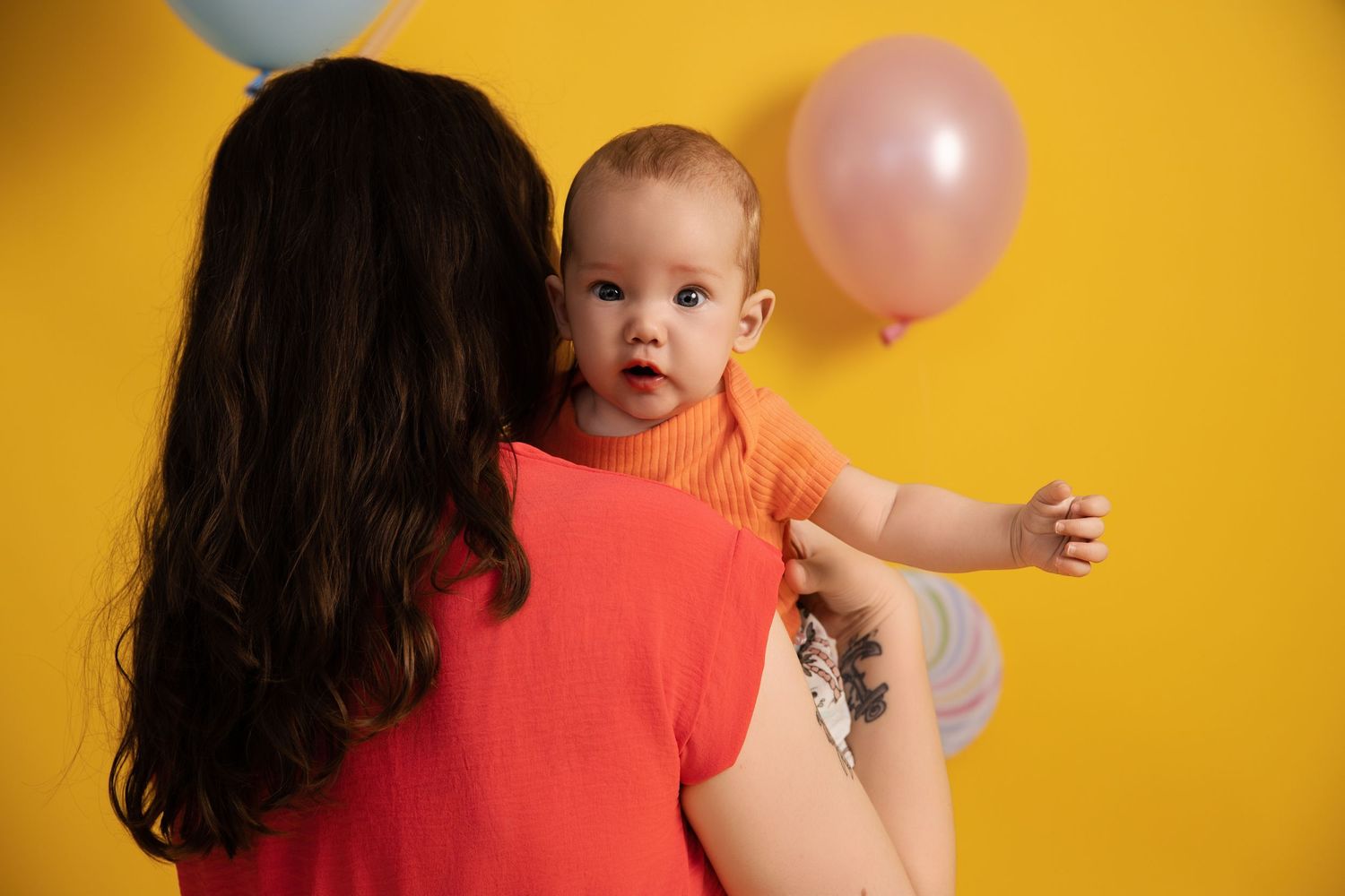 A baby in a coral shirt is held against a bright yellow background with a pink balloon visible.