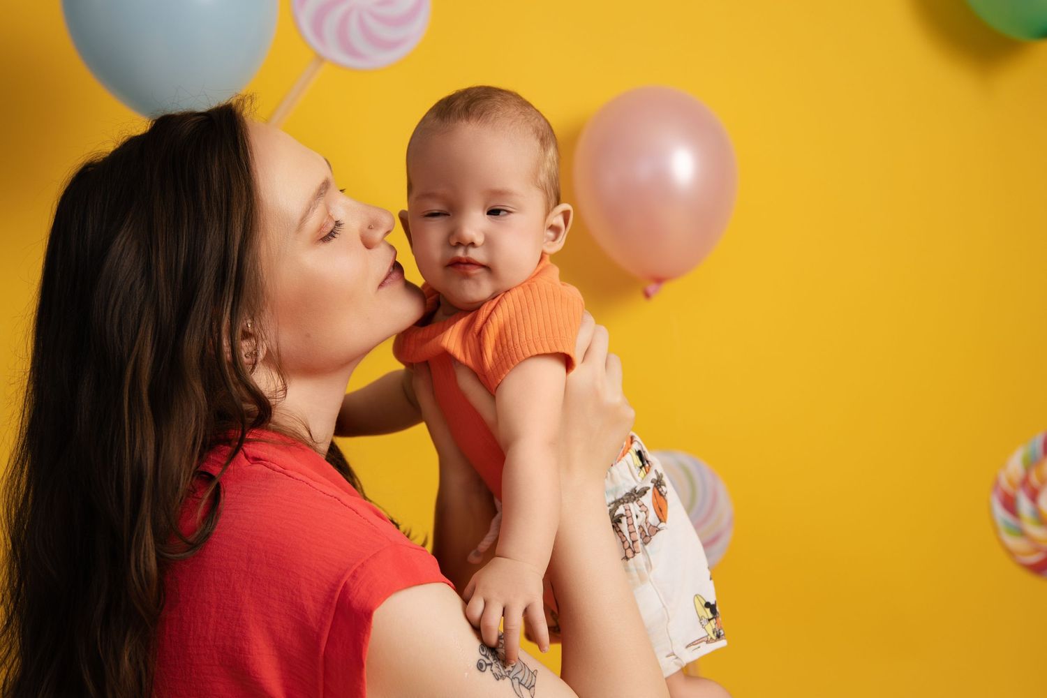 A parent and baby share a tender moment against a yellow wall with colorful party balloons in the background.