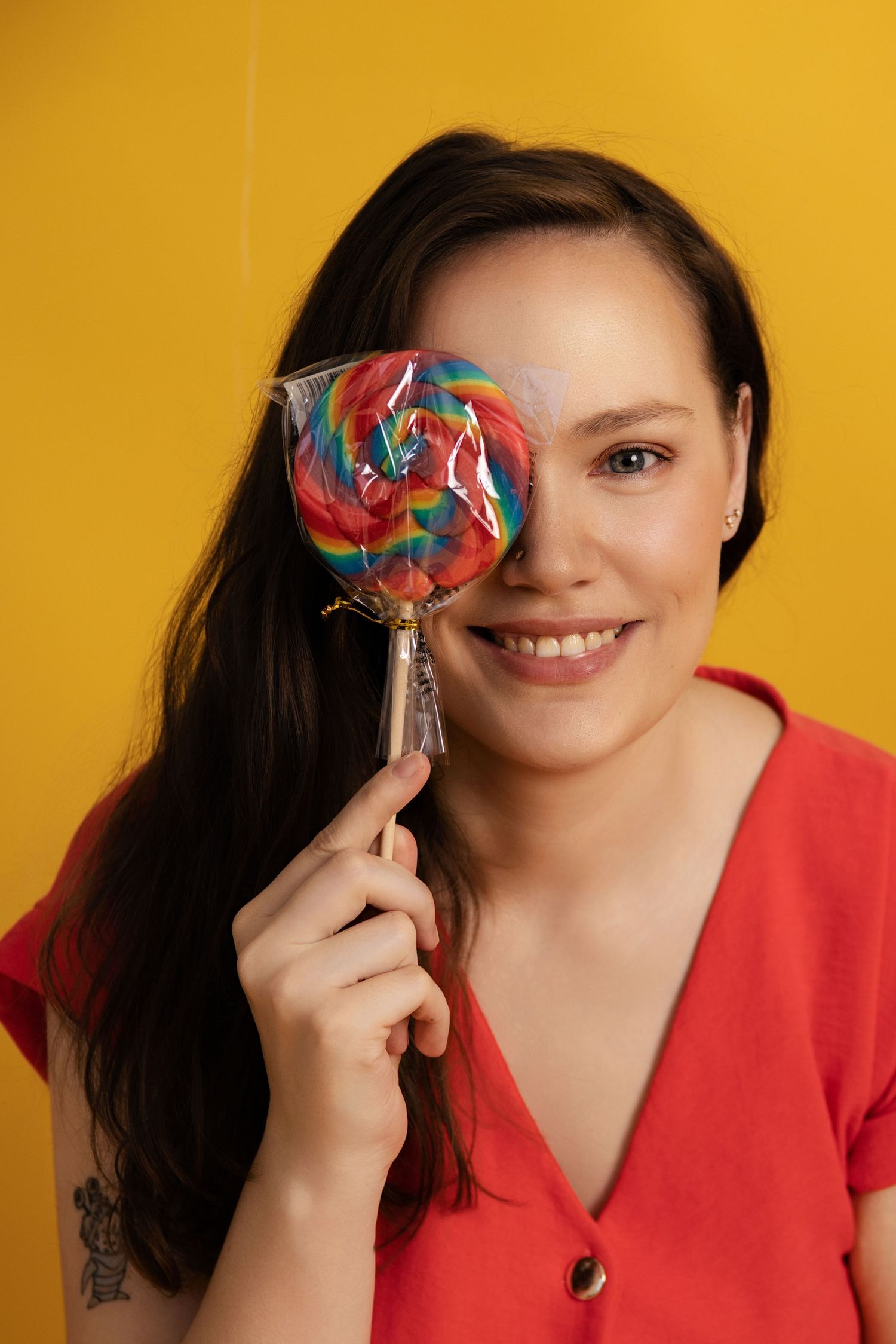 A smiling person in a red top holds a colorful rainbow lollipop against a vibrant yellow background.