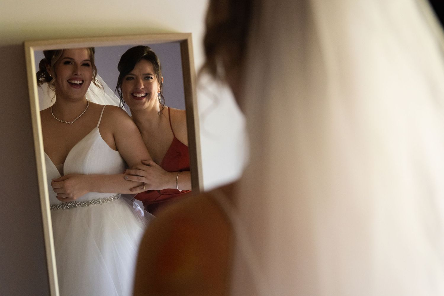 Two people in dresses pose together in front of a mirror during a wedding preparation moment.