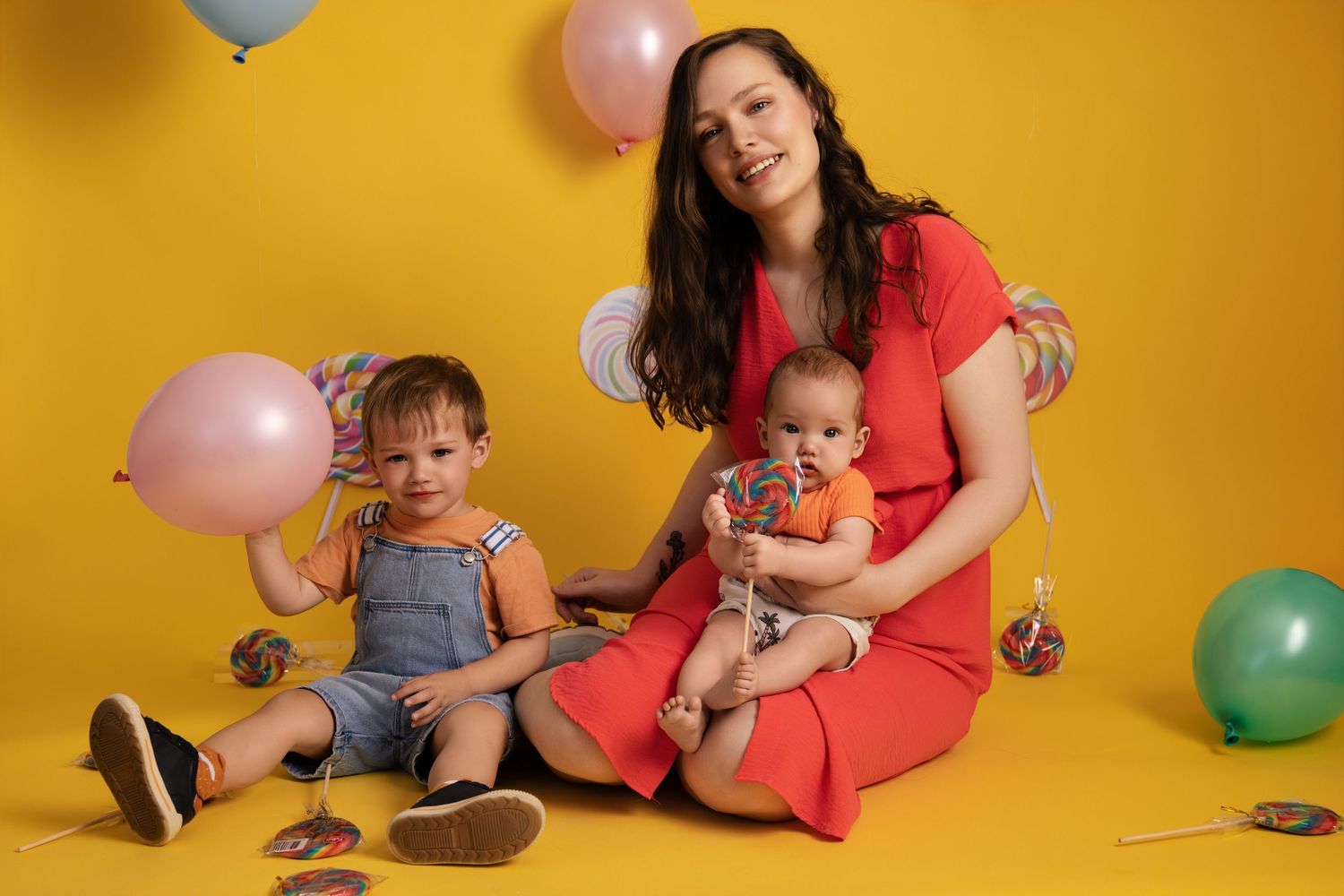 Playful family moment captured against vibrant yellow backdrop with colorful balloons and festive party decorations.
