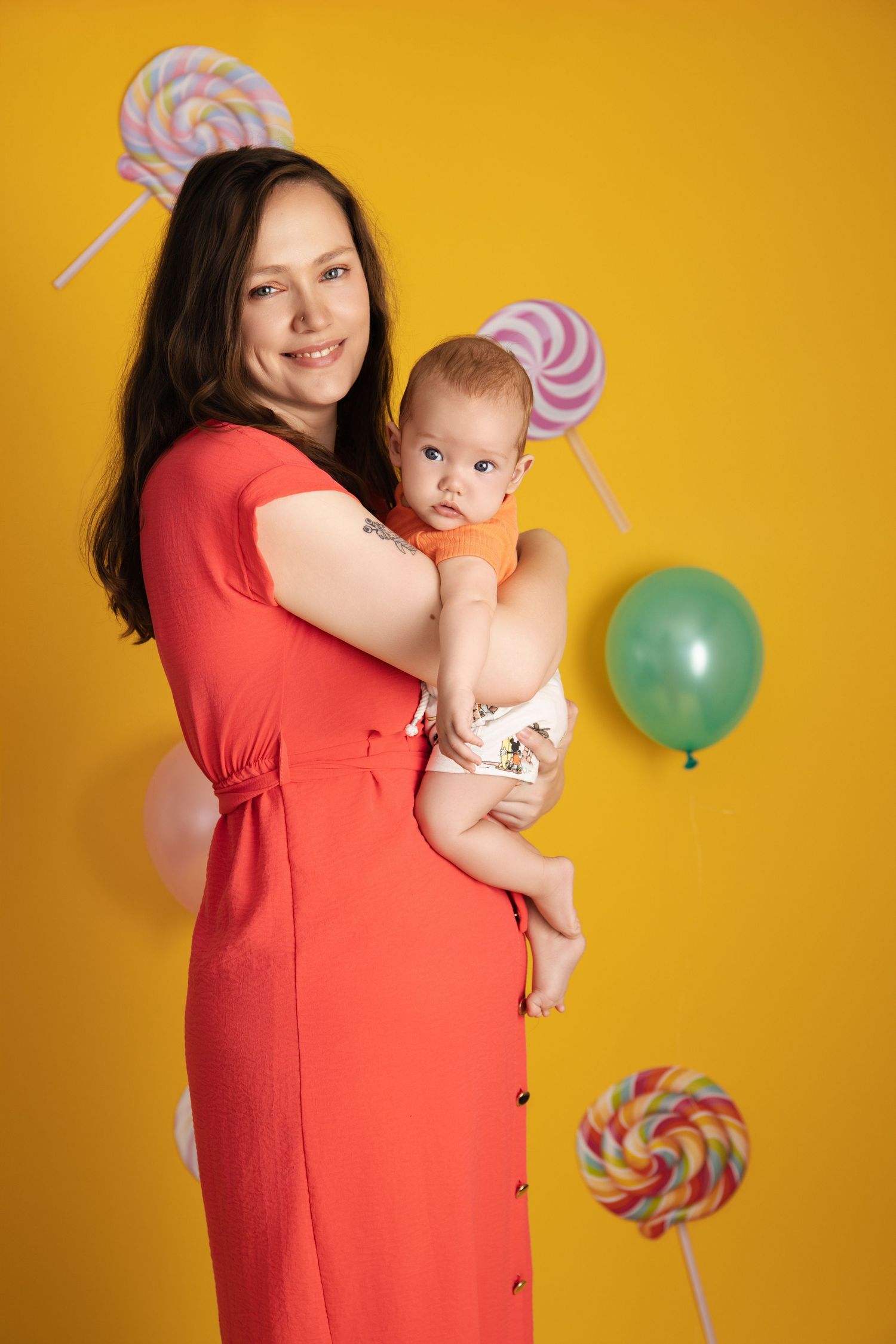 Festive scene with candy lollipop decor, green balloon, against vibrant yellow background with parent and baby.