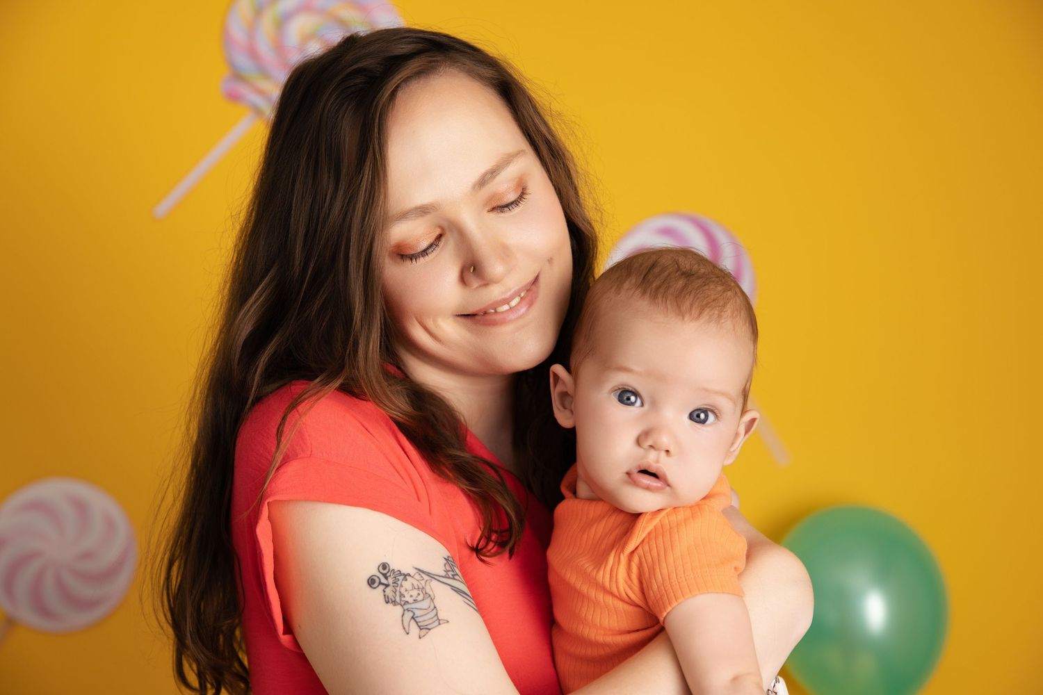 Parent and baby share a tender moment against yellow backdrop with colorful balloons and lollipop decorations.