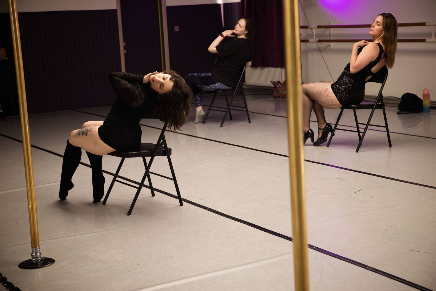 Dancers in black outfits perform a choreographed routine using chairs in a mirrored dance studio with purple lighting.