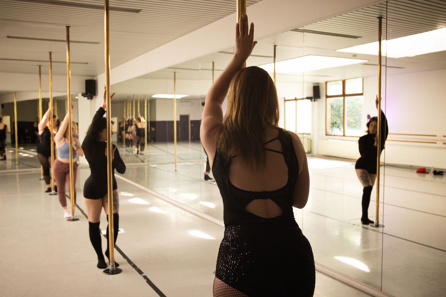 Dancers practice pole dancing moves in a mirrored studio space with bright overhead lighting.