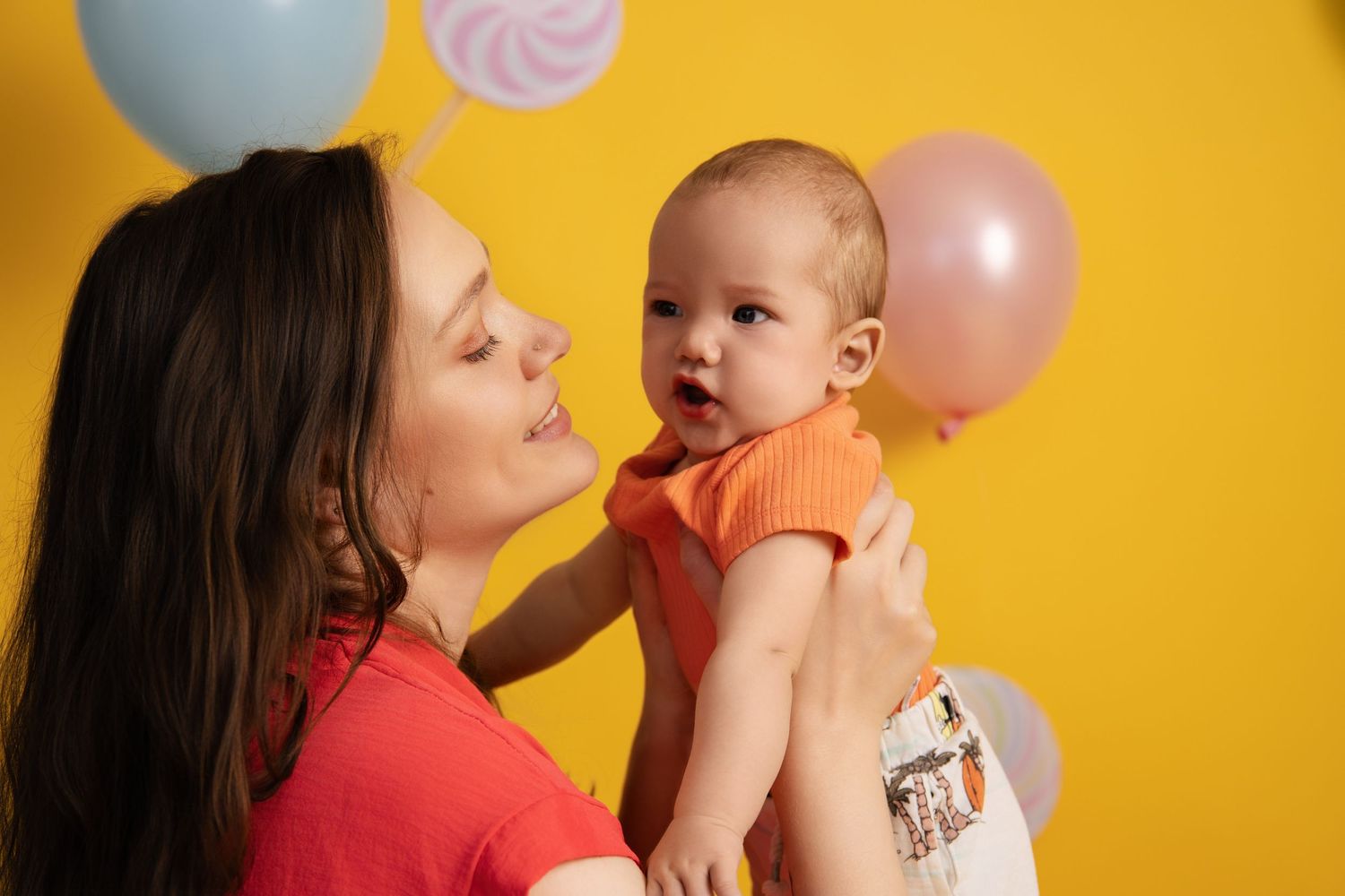 Sweet candid moment between parent and baby with balloons against vibrant yellow background.