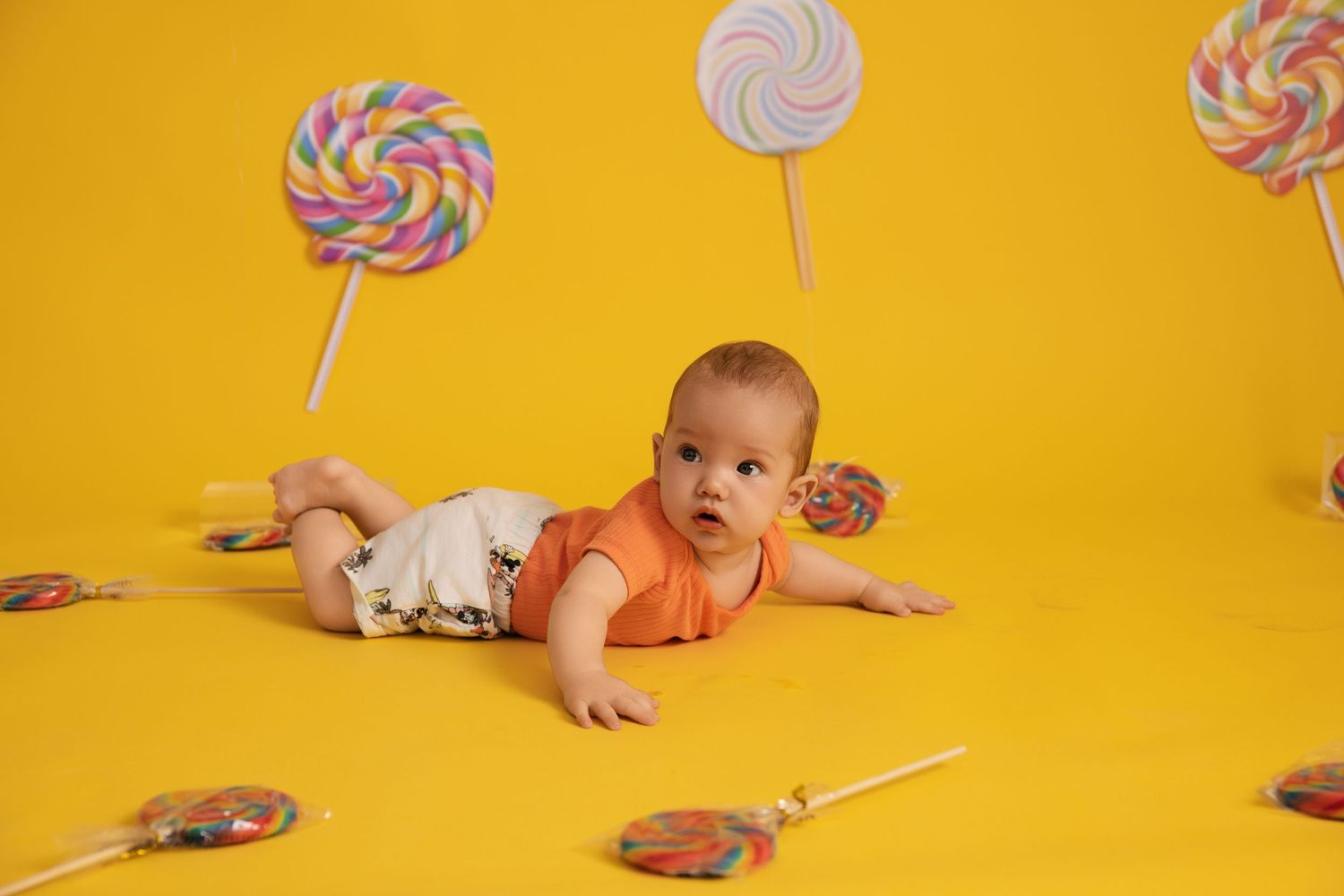 Baby in orange onesie lying on yellow background surrounded by colorful lollipop decorations.