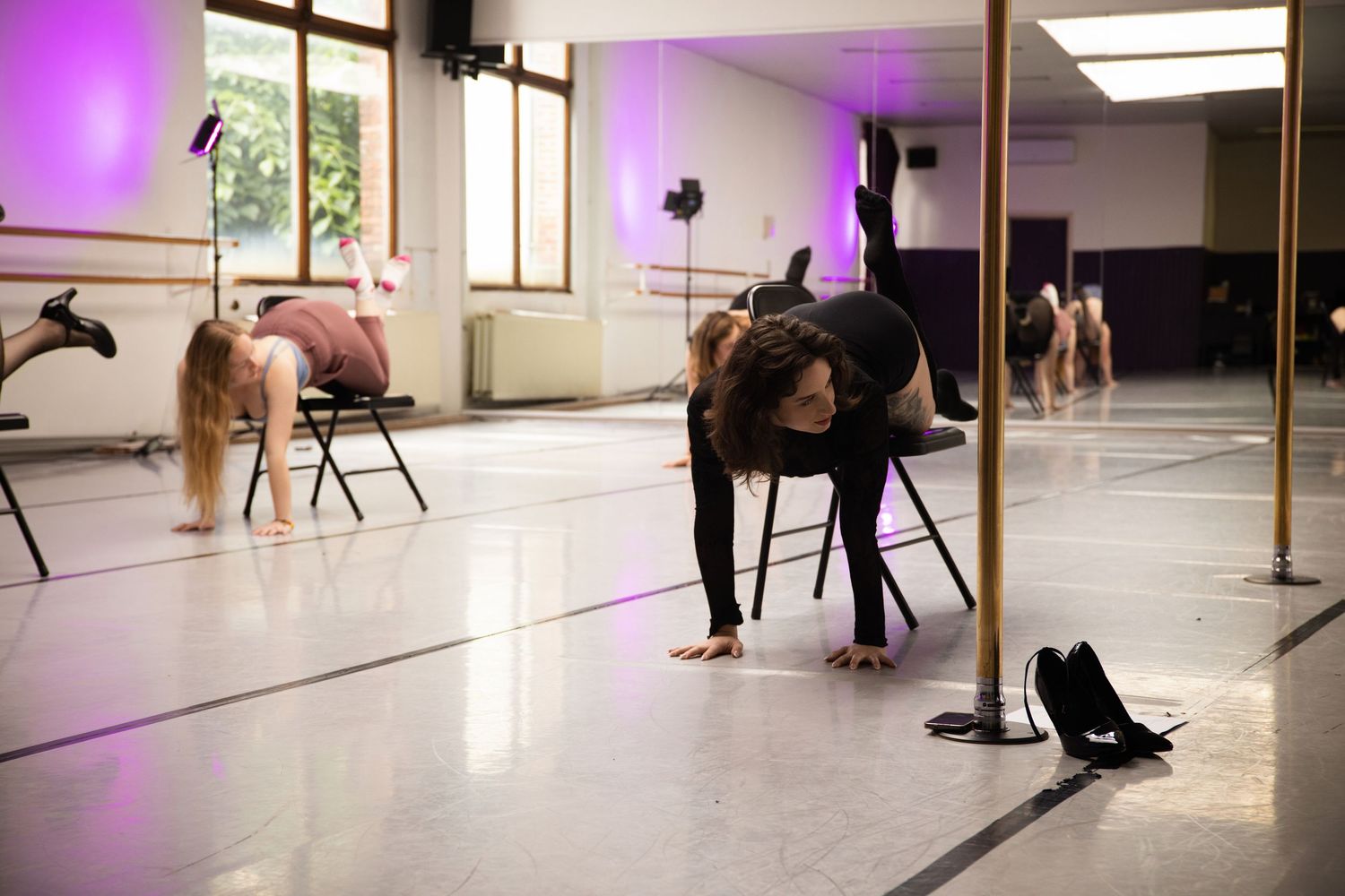 Dancers warm up and stretch in a bright dance studio with purple lighting and ballet barres along mirrored walls.