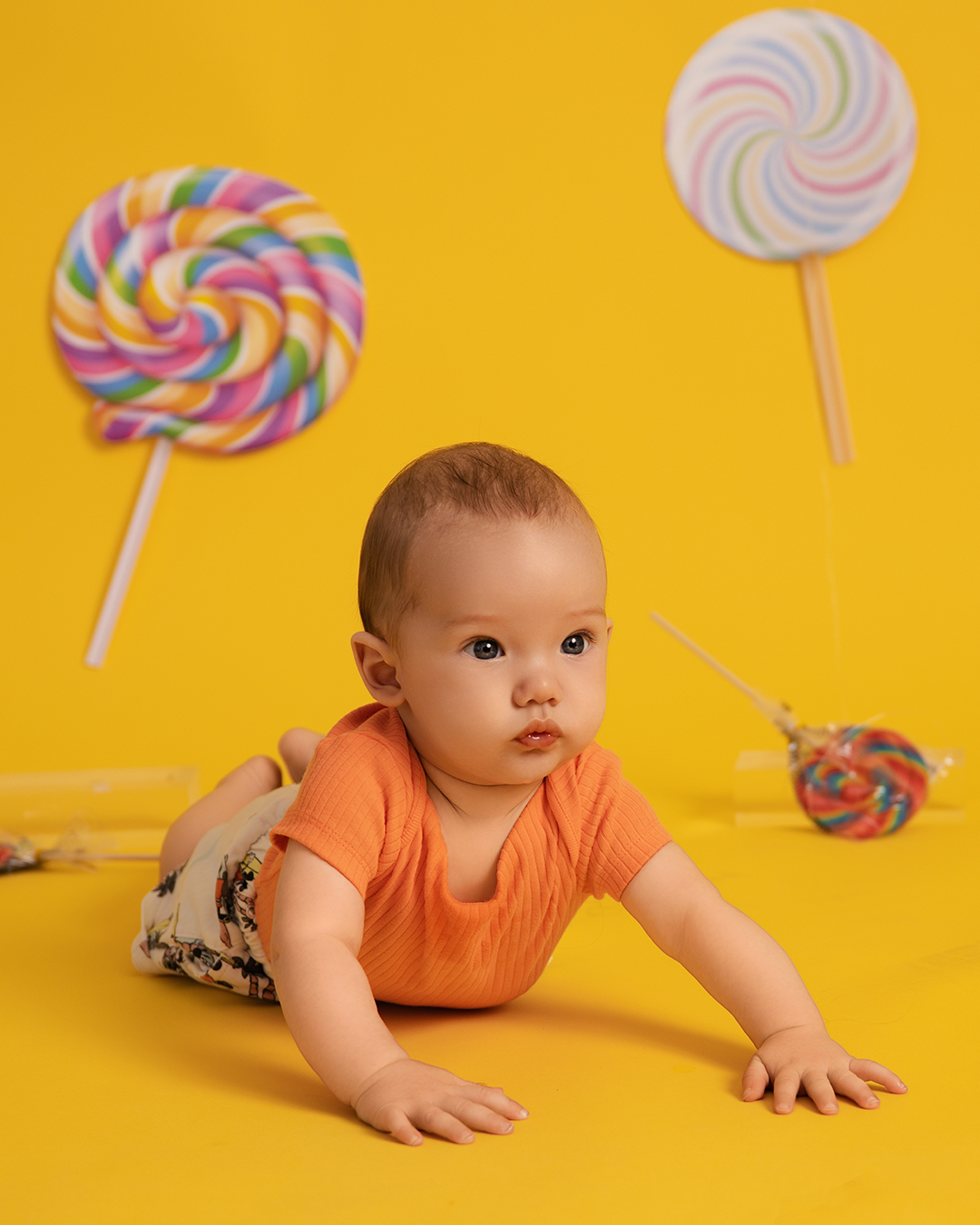 Baby in orange outfit crawls across yellow backdrop with colorful lollipop decorations in progressive motion.