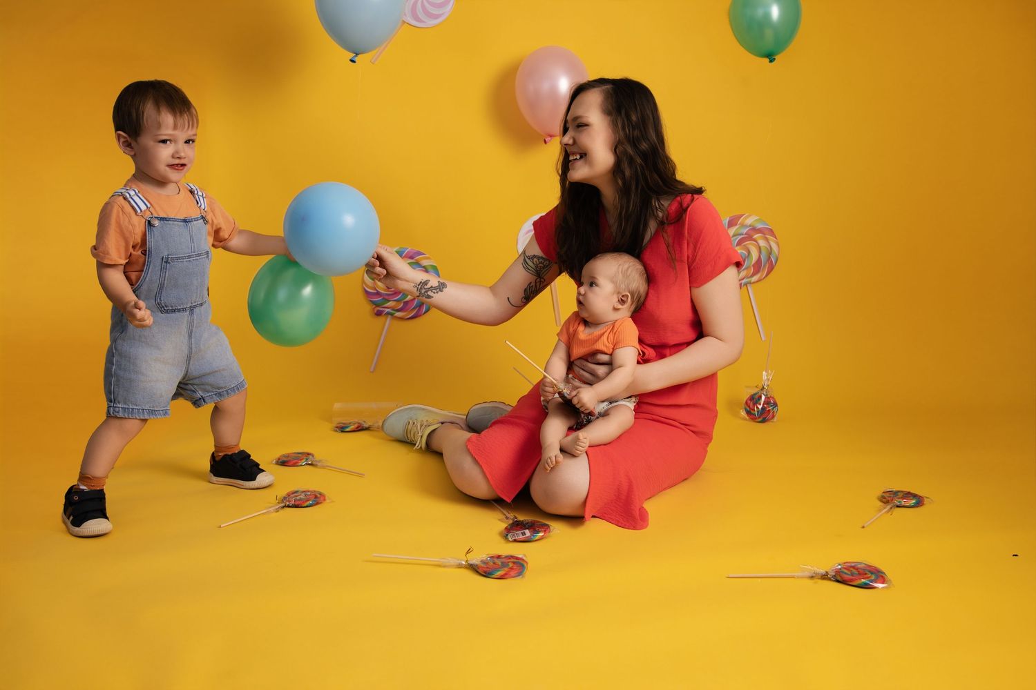 Parent and toddler play with colorful balloons against a vibrant yellow studio backdrop in a series of candid moments.