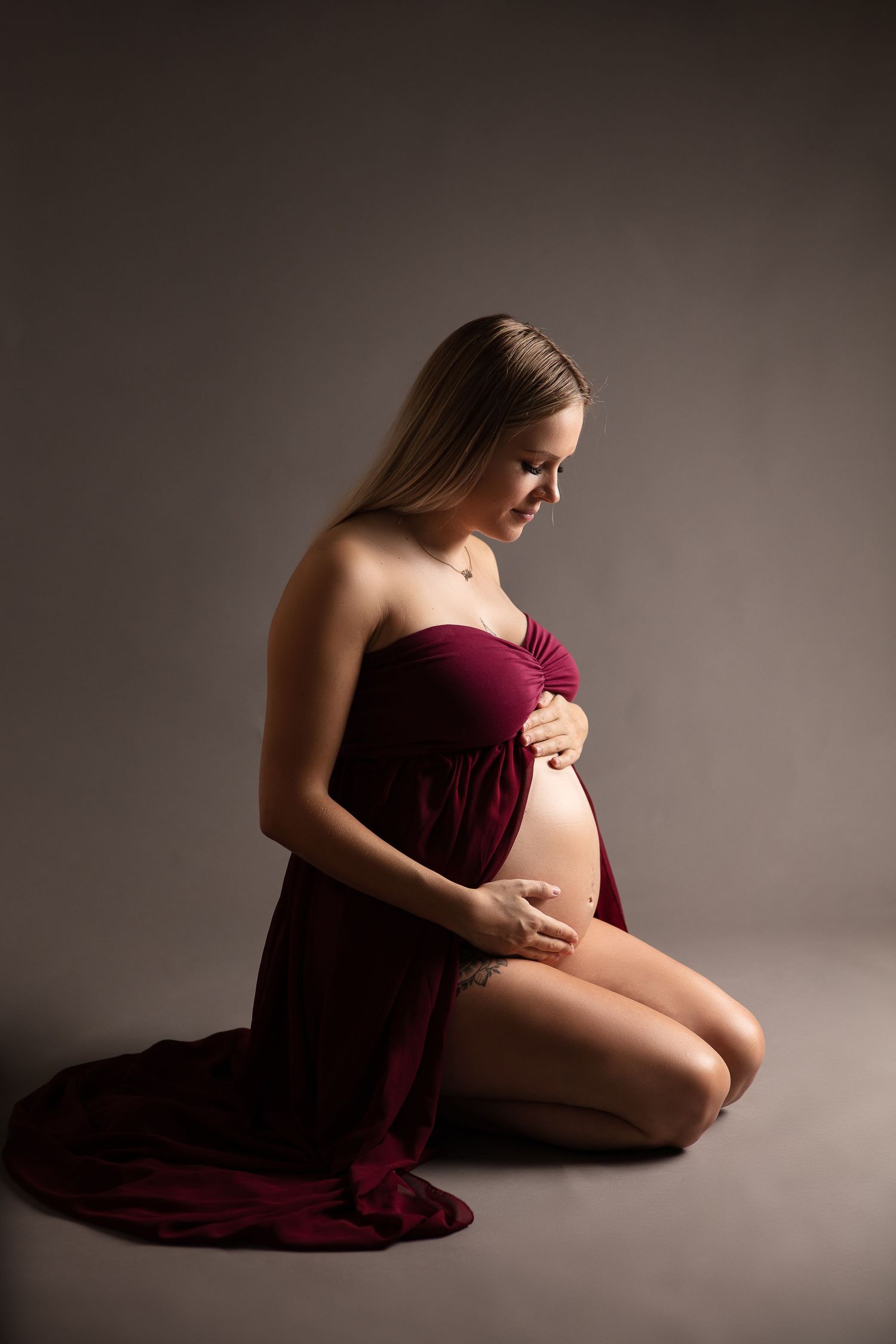 Artistic maternity portrait featuring an expectant mother in burgundy fabric kneeling gracefully against a dark background.