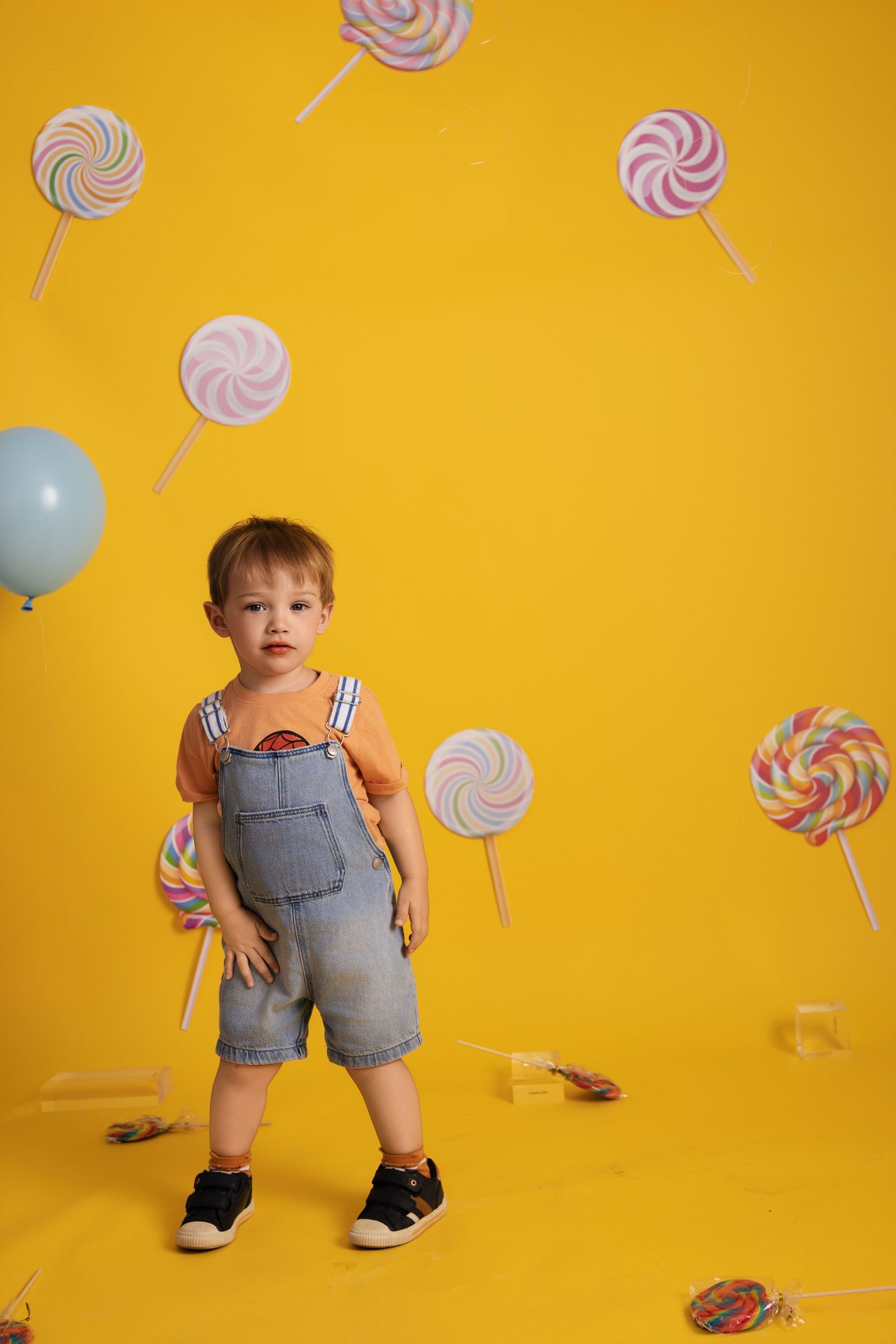 Whimsical yellow backdrop with floating lollipop decorations and blue balloon, featuring young toddler in denim overalls.