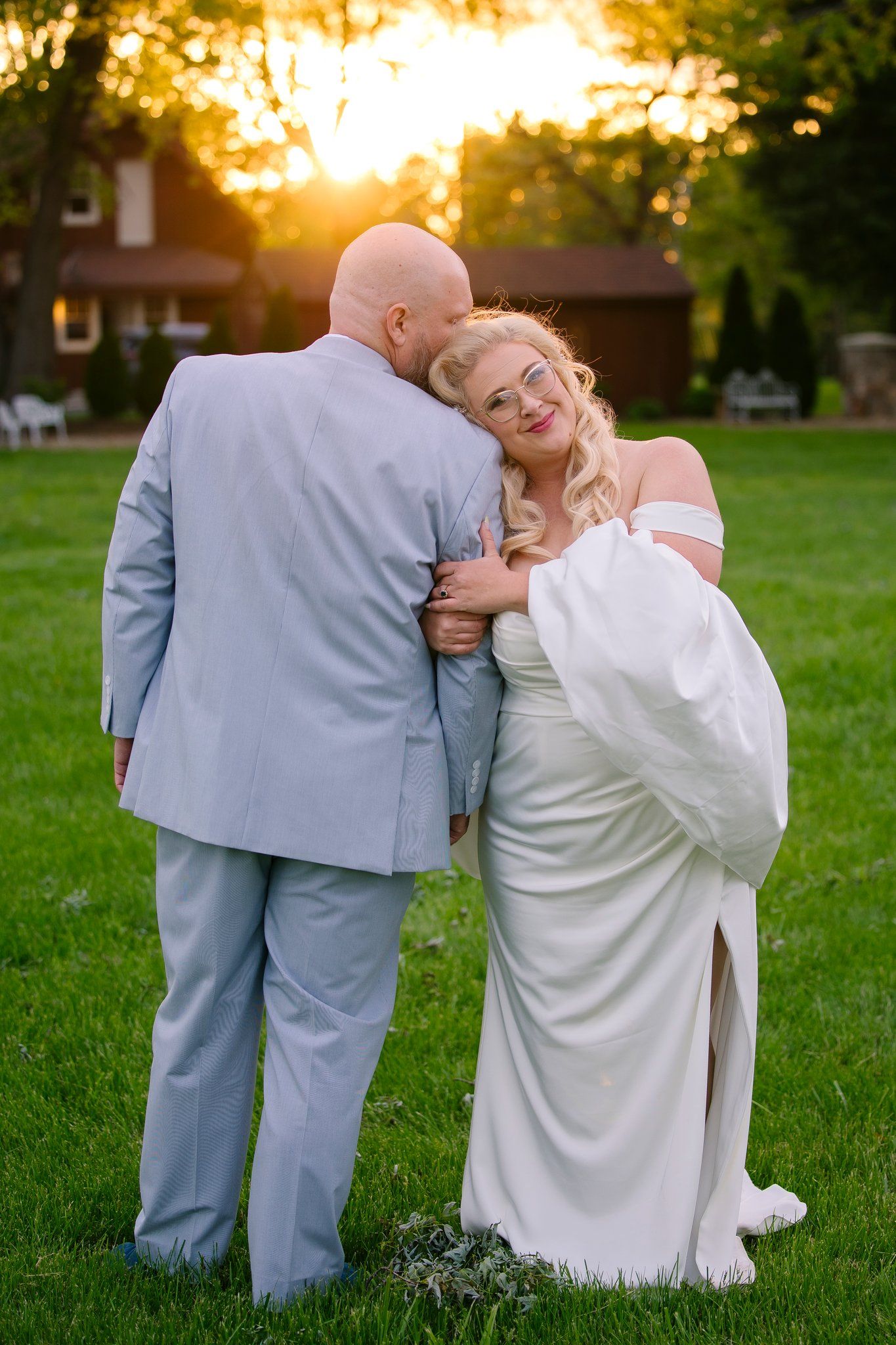 A romantic golden hour wedding photo of a couple in gray suit and white dress posing on grass at sunset.