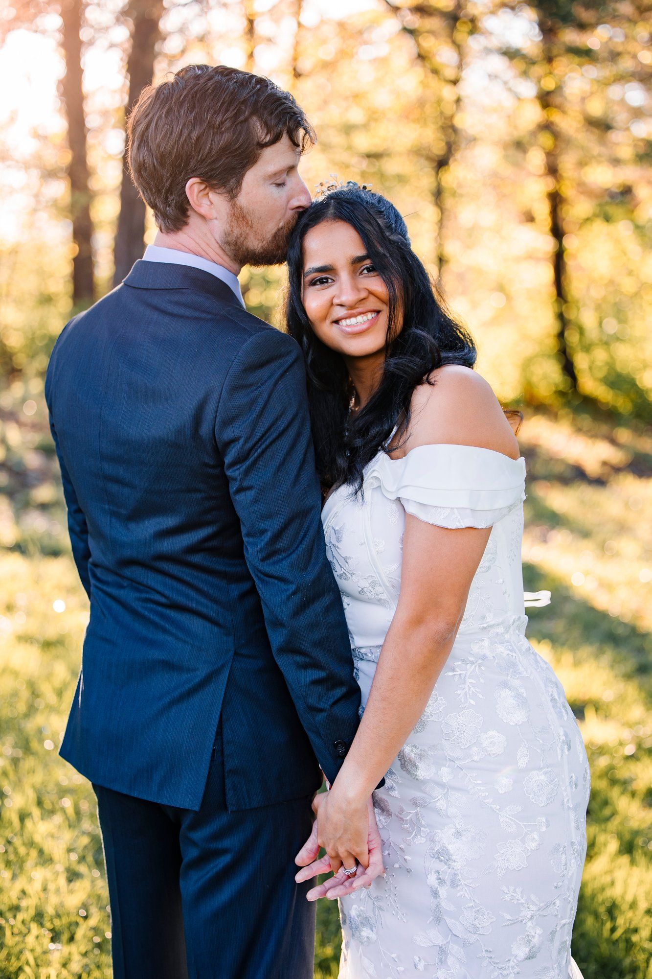 A couple in wedding attire holds hands while standing in a sun-dappled forest during golden hour.
