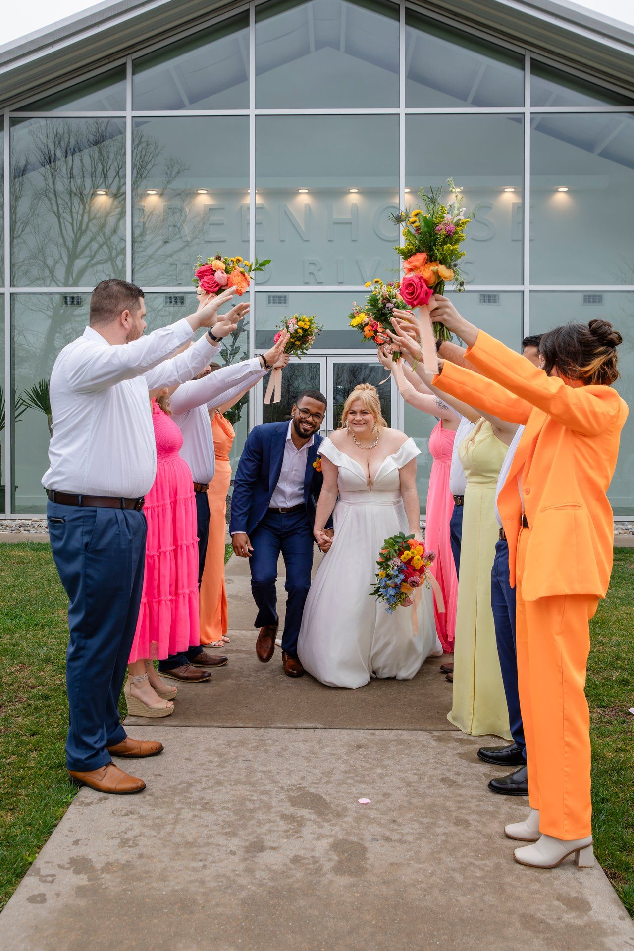 Wedding guests in bright attire form a celebratory arch with bouquets for the newlyweds.