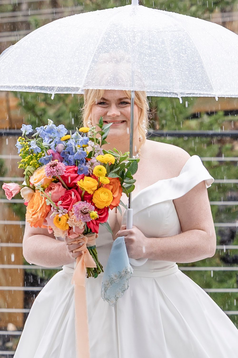 A cheerful bride in a white wedding dress holds a vibrant multicolored bouquet while standing under a clear umbrella in the rain.