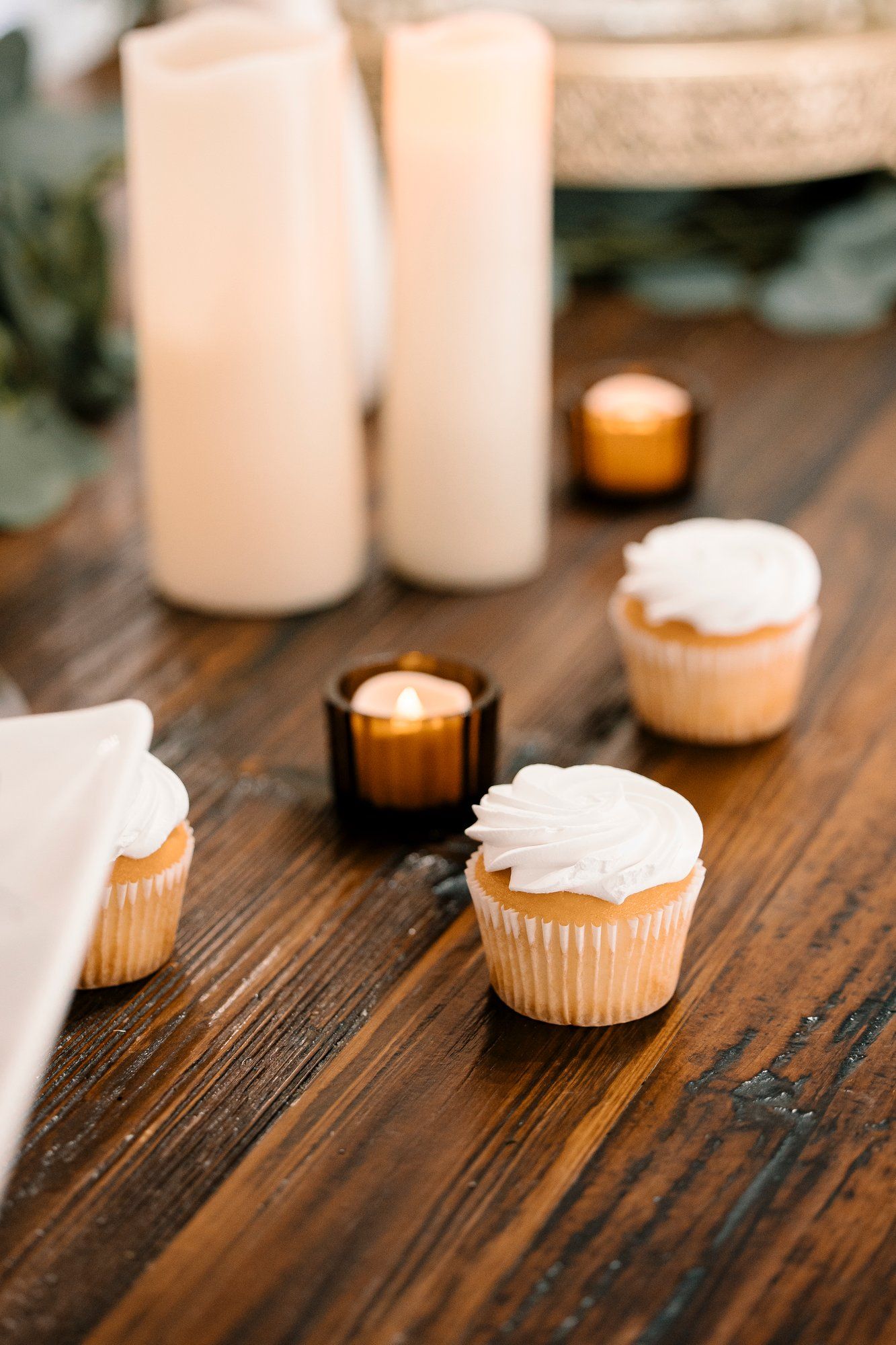 Vanilla cupcakes with white frosting and glowing candles on rustic wooden table with tall white pillar candles in background.