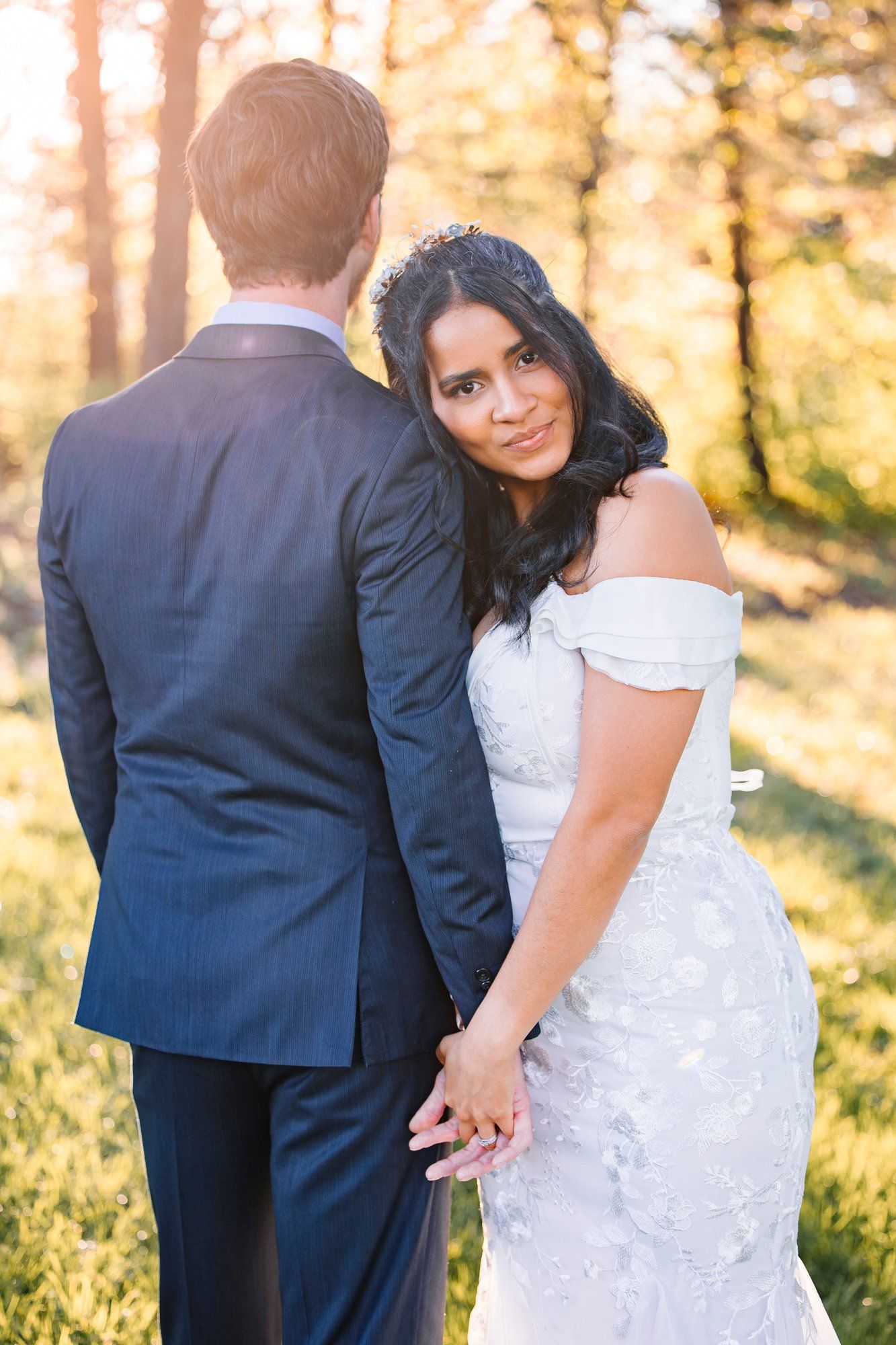 A newlywed couple stands close together in a golden sunlit forest setting wearing wedding attire.