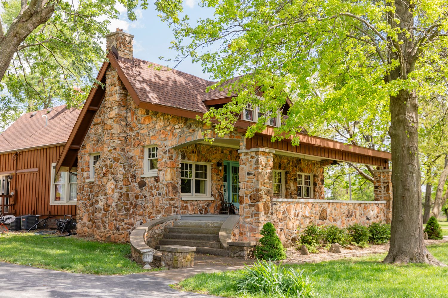 A charming stone cottage with a rustic porch and brown roof surrounded by green trees in a peaceful setting.