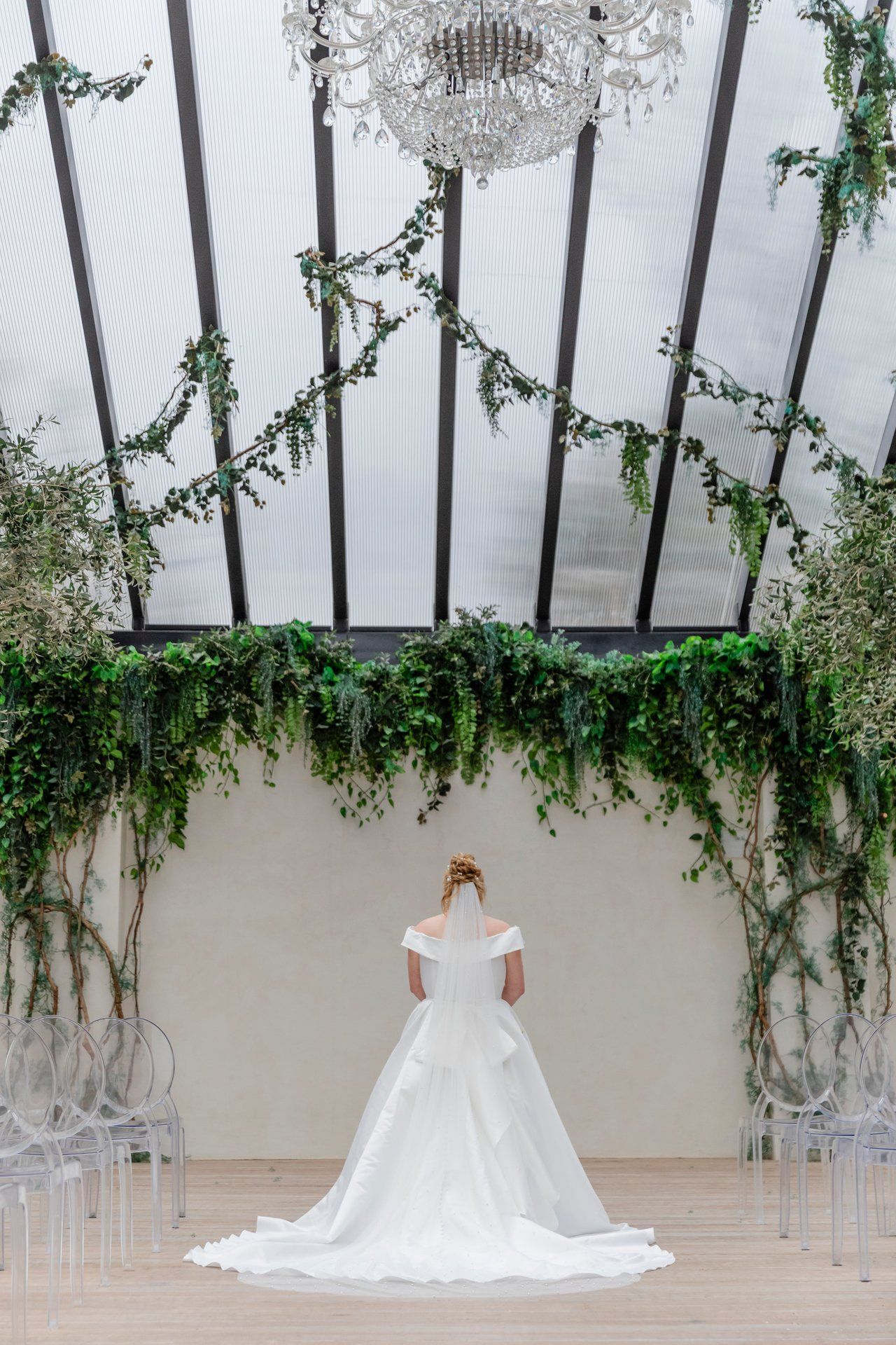 A white wedding gown photographed from behind under a crystal chandelier and greenery-adorned ceiling.