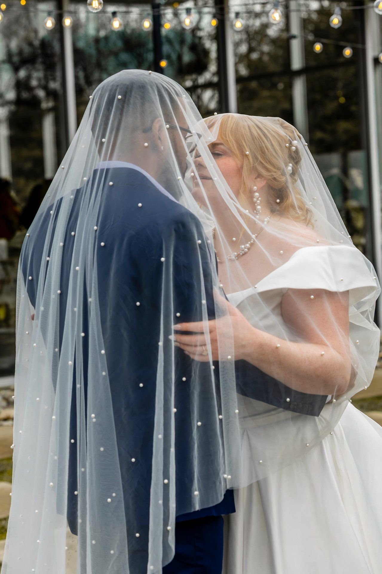 A romantic wedding moment captured through a sheer veil with twinkling string lights in the background.