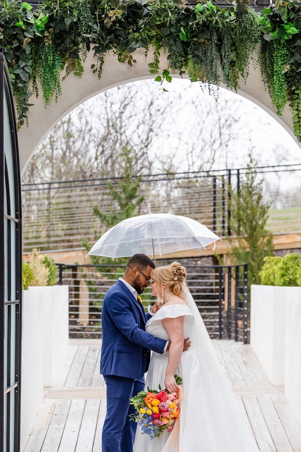 Newlyweds share a moment under a clear umbrella in a garden setting with hanging greenery.