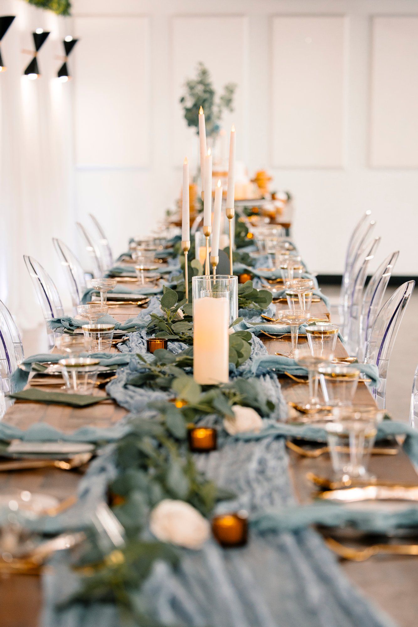 Long banquet table decorated with eucalyptus garland, white candles, and modern place settings in a bright white venue.