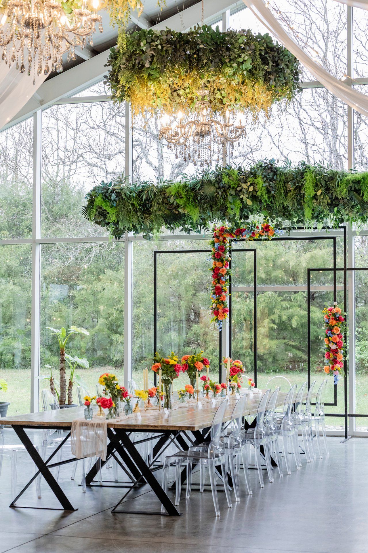 Modern glass wedding venue with hanging floral chandeliers, geometric frames, and a long dining table setup with ghost chairs.