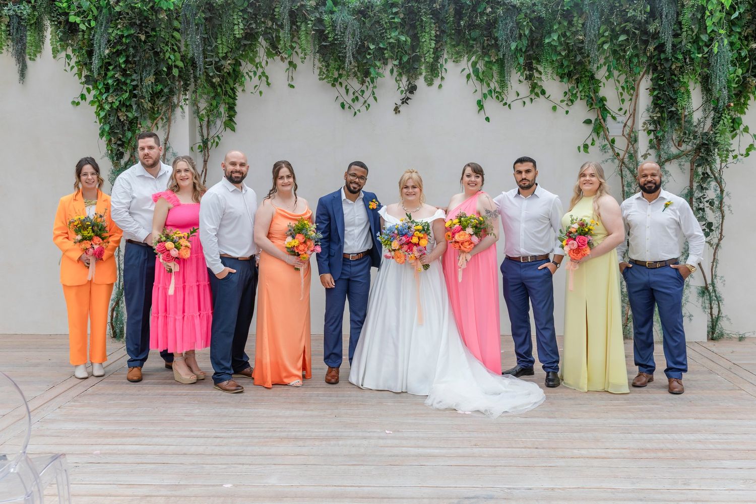 Wedding party poses under hanging greenery with colorful bridesmaid dresses.