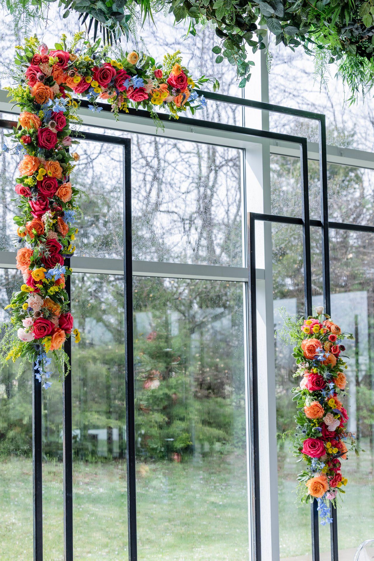 Modern black metal wedding arch decorated with vibrant red and orange flowers against glass windows.