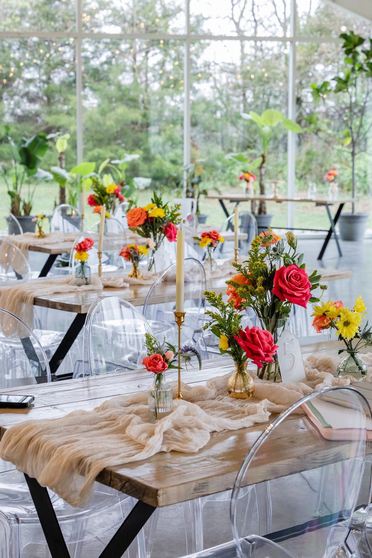 Modern rustic tablescape with bright floral centerpieces, ghost chairs, and wooden tables in a light-filled greenhouse setting.