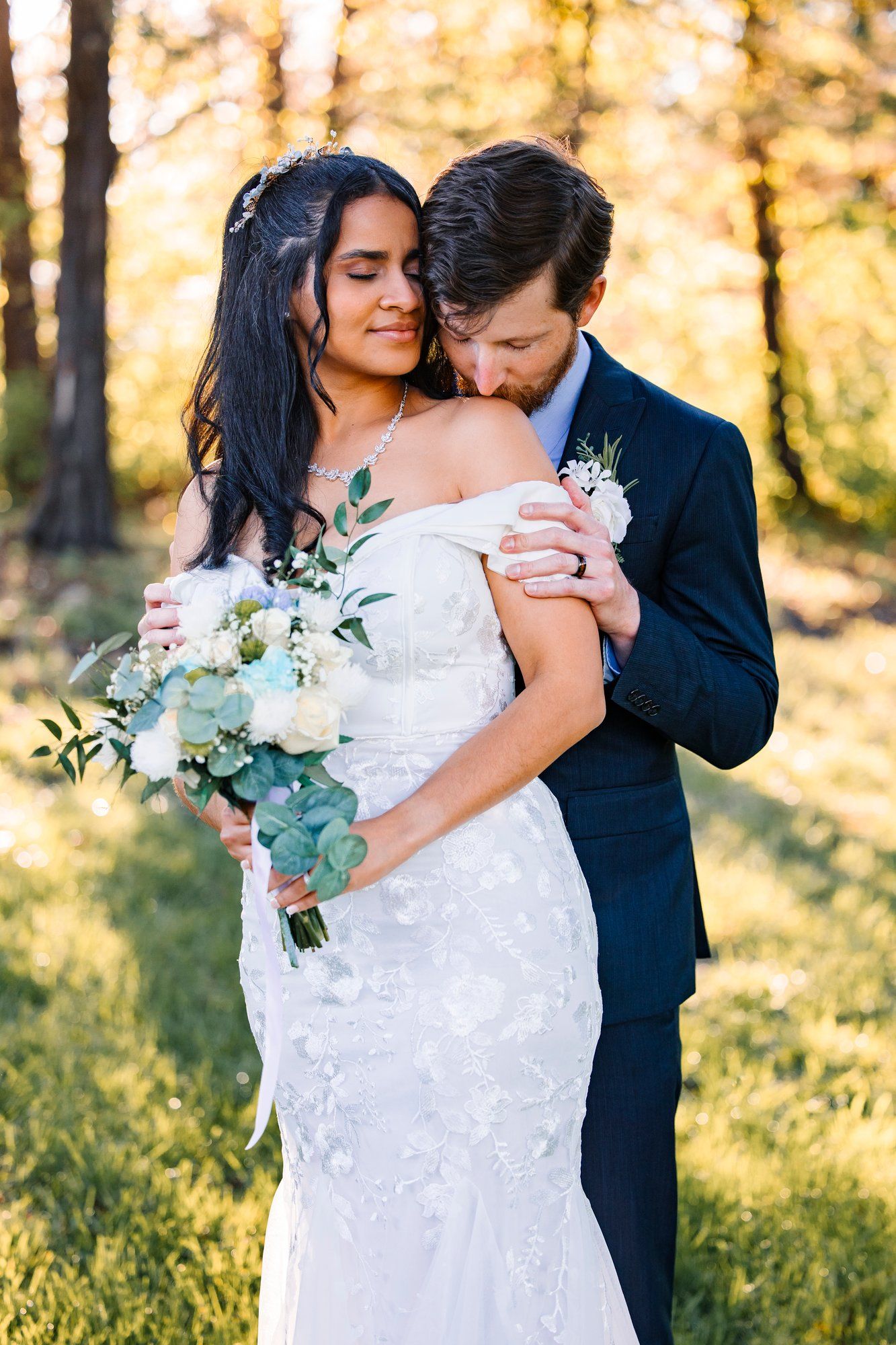 Romantic newlyweds embrace in golden sunlit forest setting with bride holding eucalyptus and white flower bouquet.