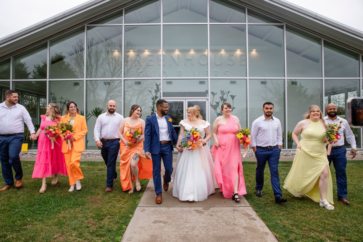 Wedding party poses in colorful pink and orange attire outside a modern glass building with string lights.