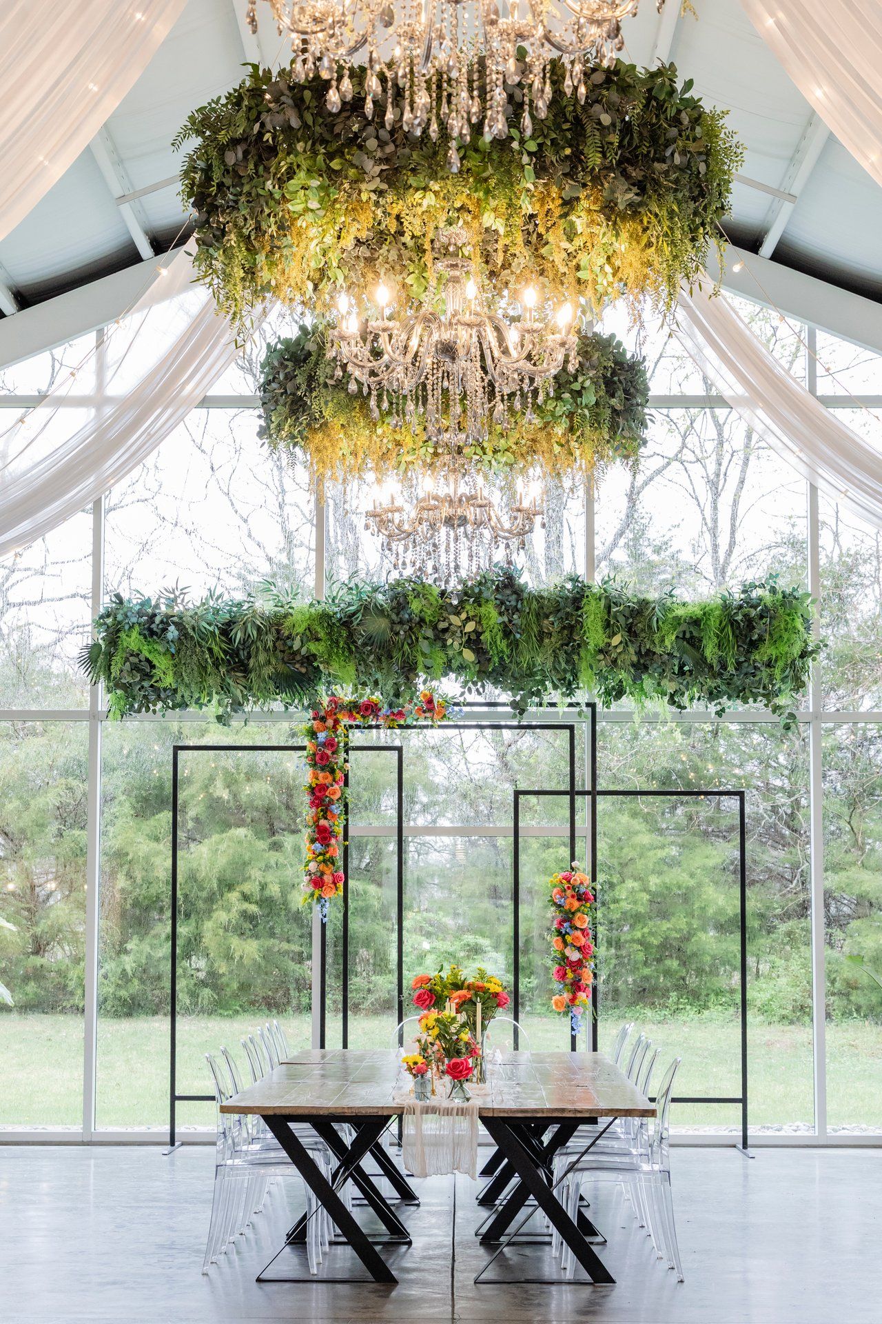 Elegant greenhouse wedding setup with tiered crystal chandeliers adorned with greenery and modern black metal arches.