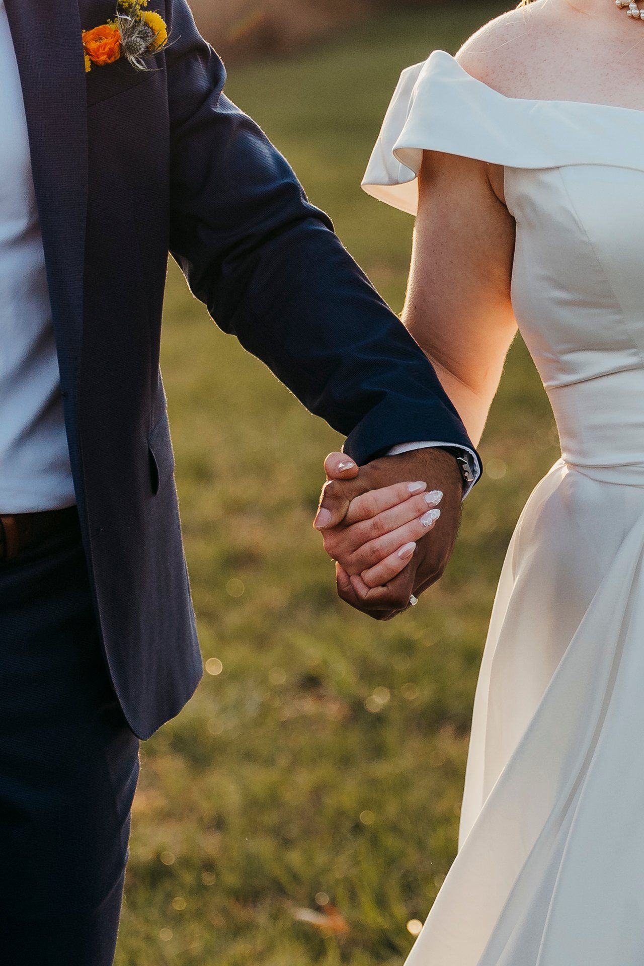 Close-up of newlyweds holding hands, showing wedding attire details and orange boutonniere.