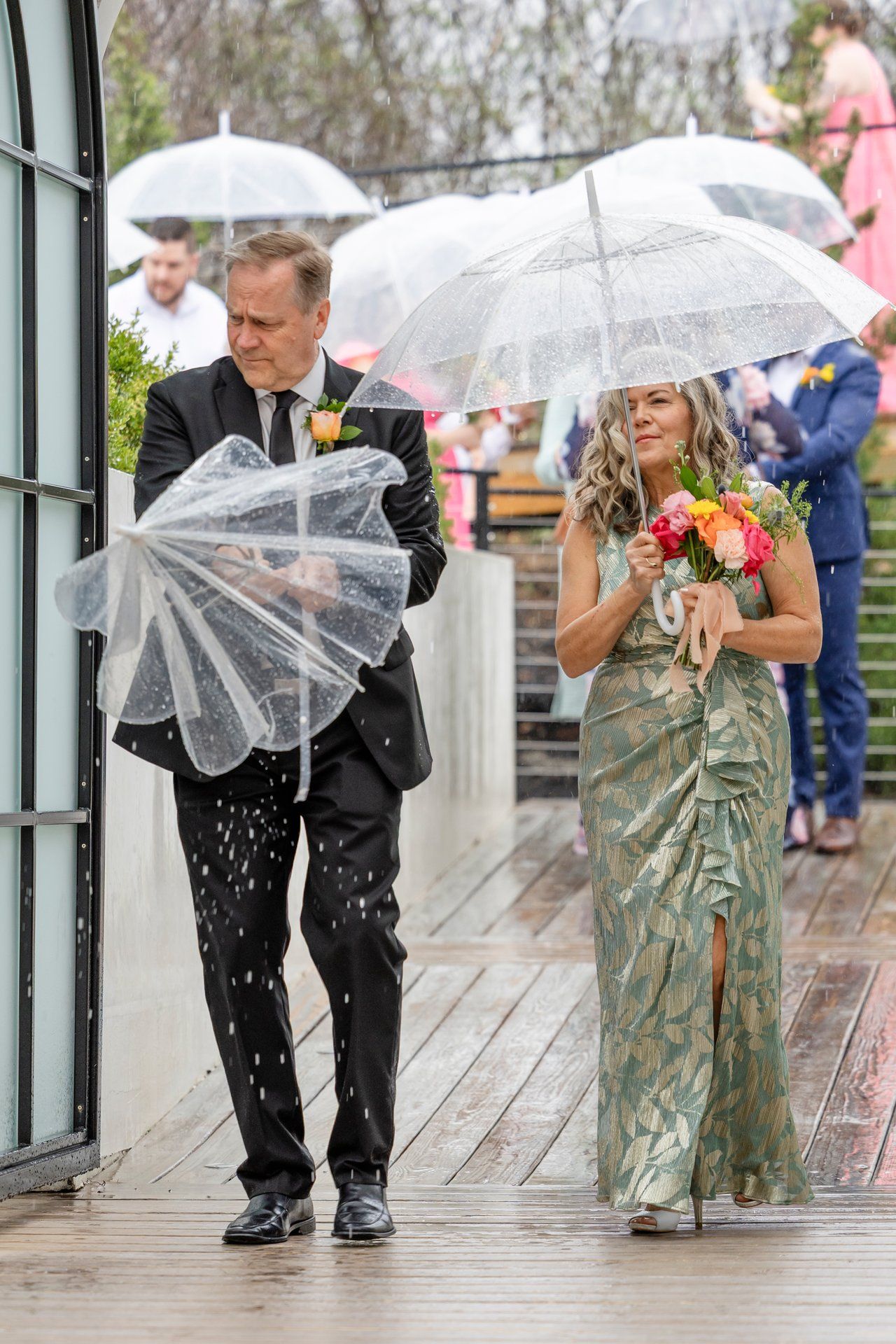Wedding party members walk under clear umbrellas on a rainy day, with a woman in a green dress holding a bright bouquet.