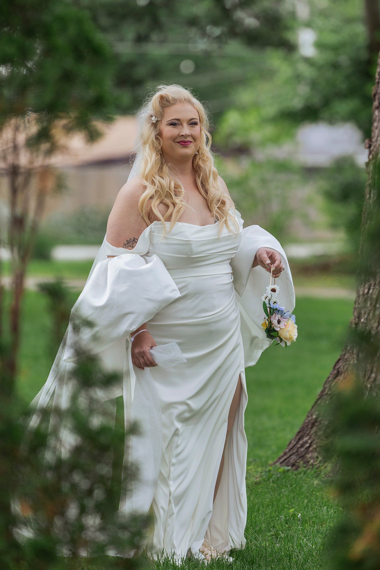 A bride in an elegant white wedding dress twirls playfully outdoors in a lush green garden setting.